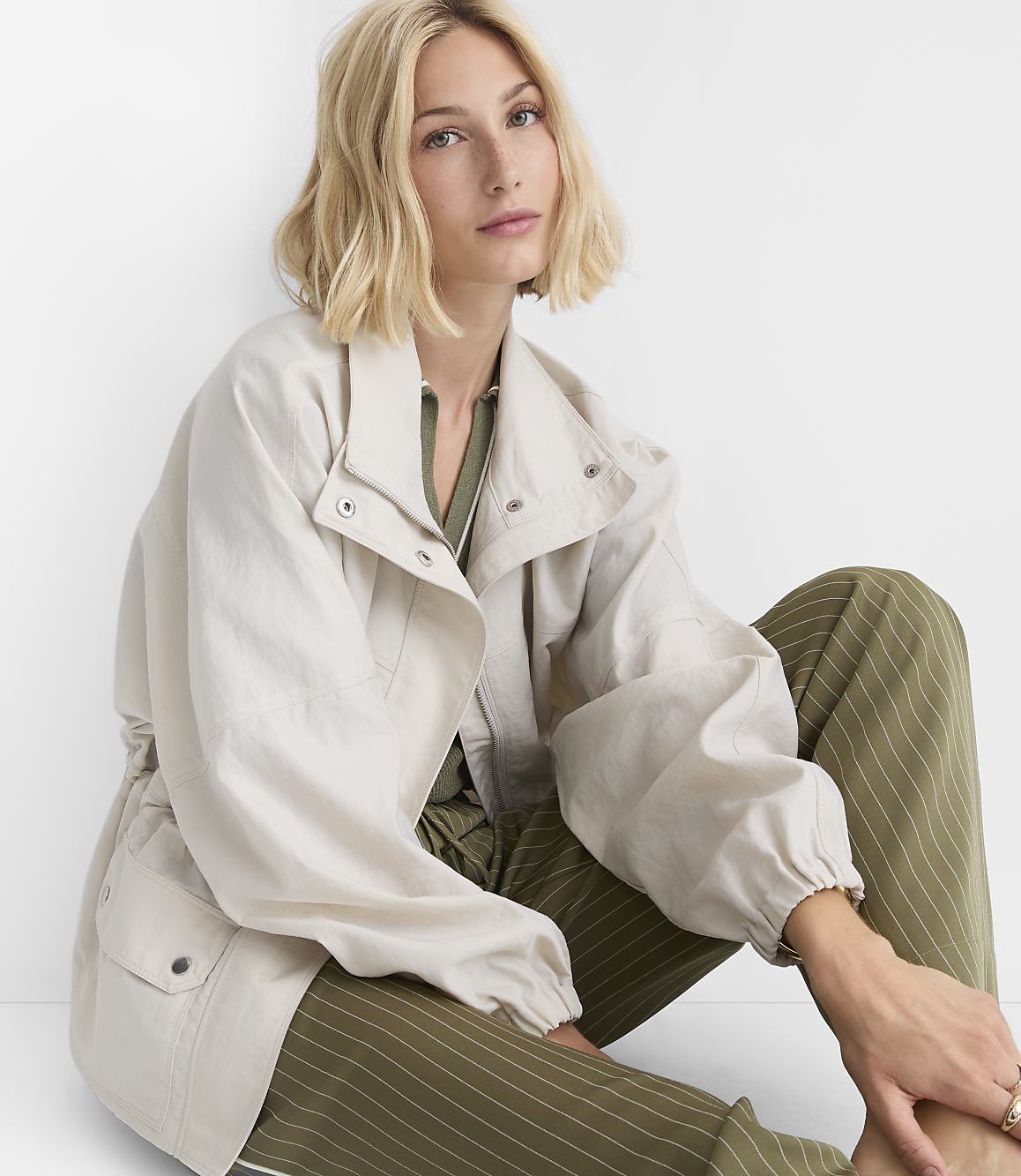 A blonde woman in a light jacket and green striped pants sits against a plain white background.