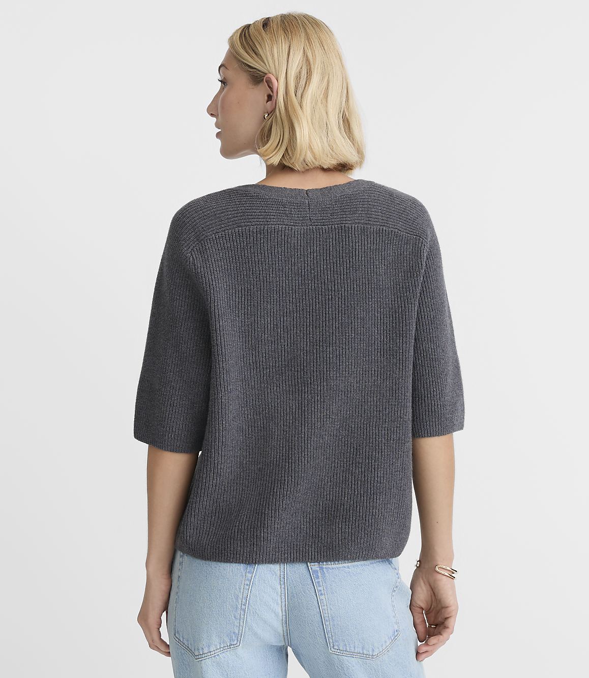 A woman in a gray sweater and light blue jeans stands against a plain white background.