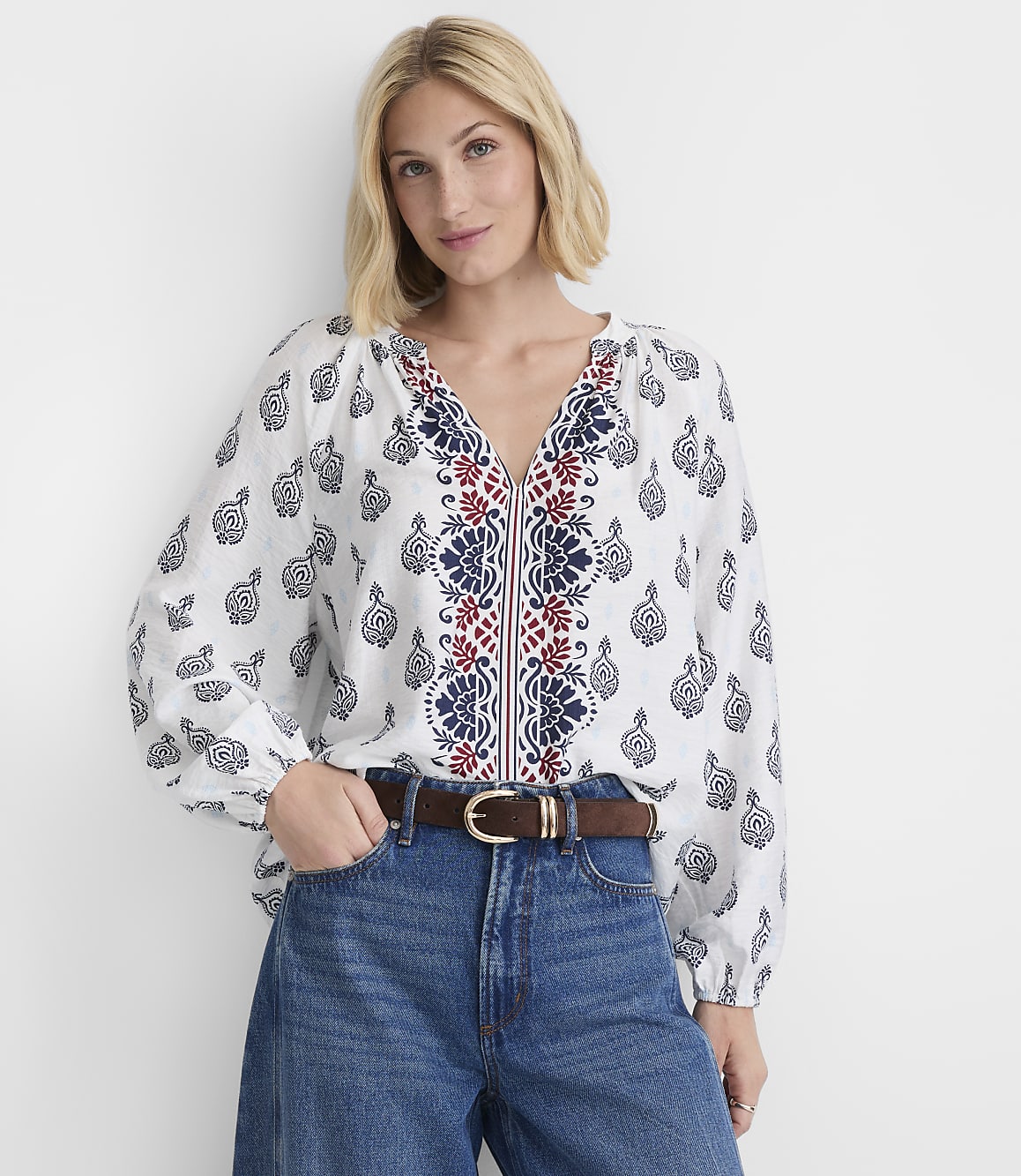 A woman in a white embroidered blouse and blue jeans stands against a plain white background.