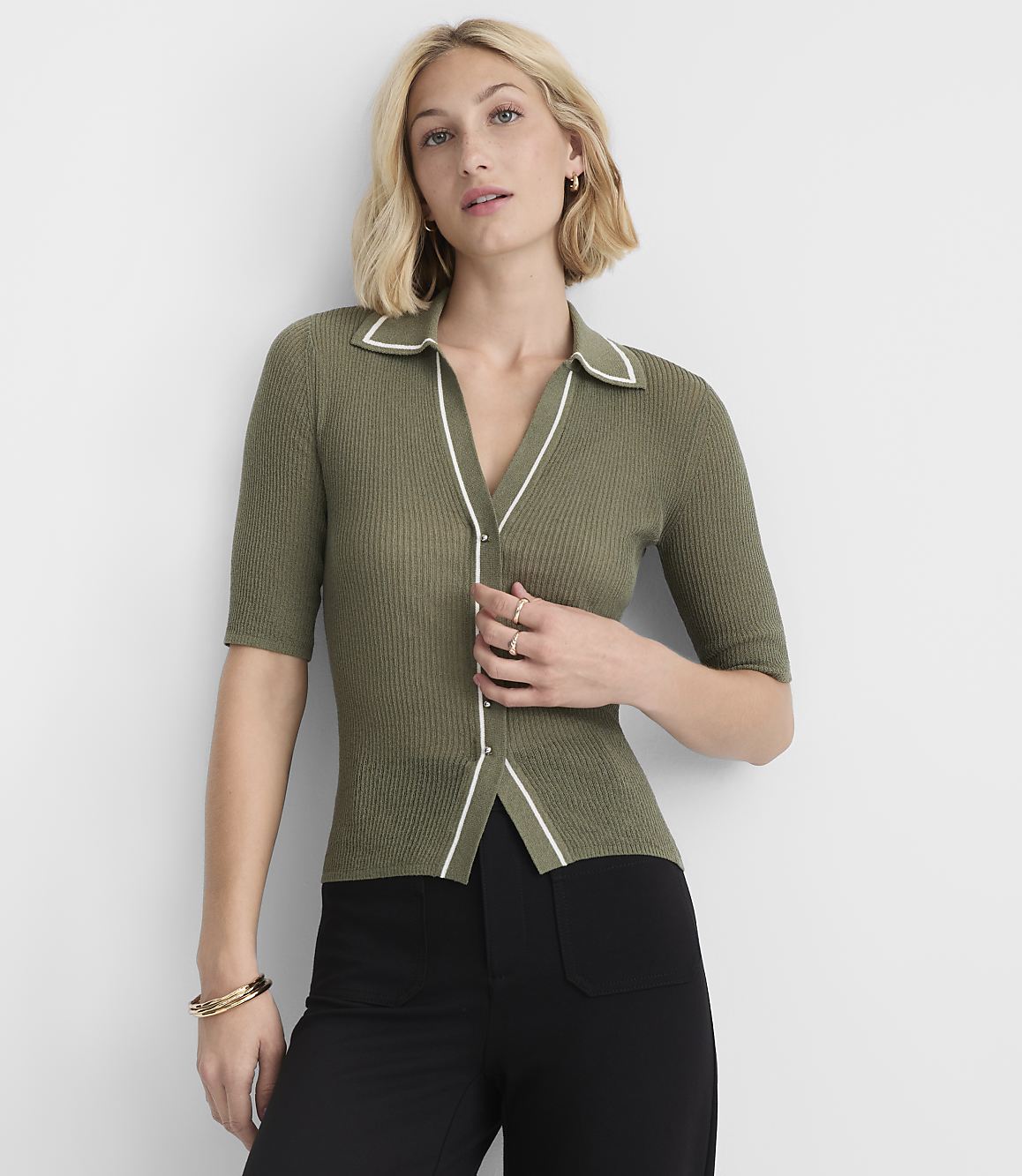 A woman in a green collared shirt and black pants stands against a plain white background.