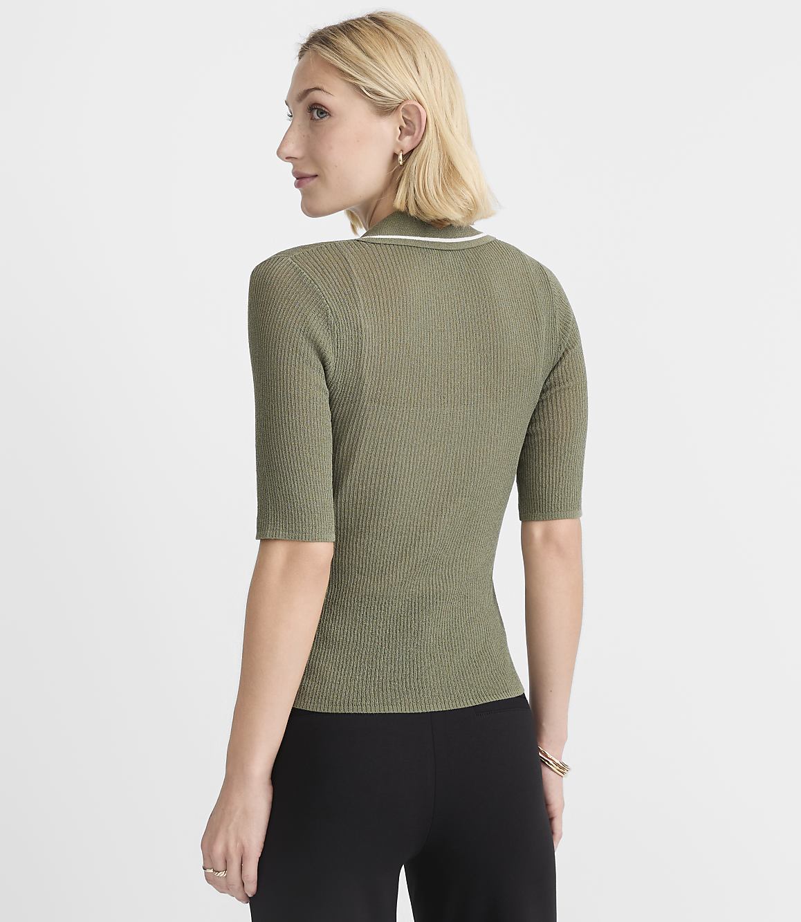 A woman in a green ribbed shirt and black pants stands against a plain white background.