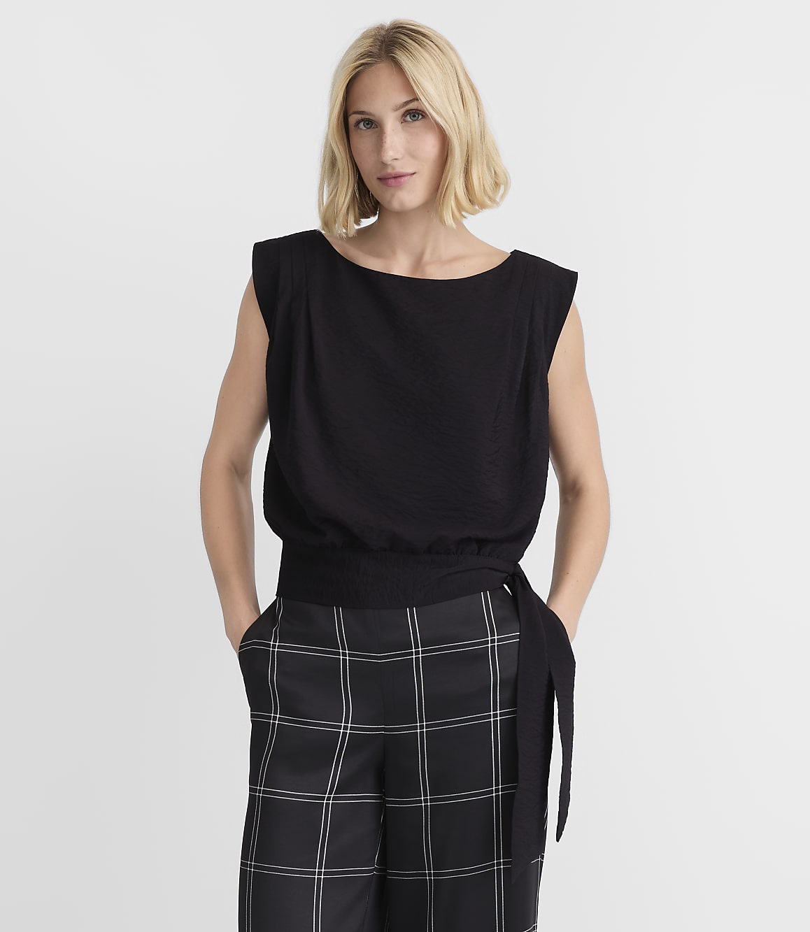 A blonde woman in a black top and plaid pants stands against a plain white background.