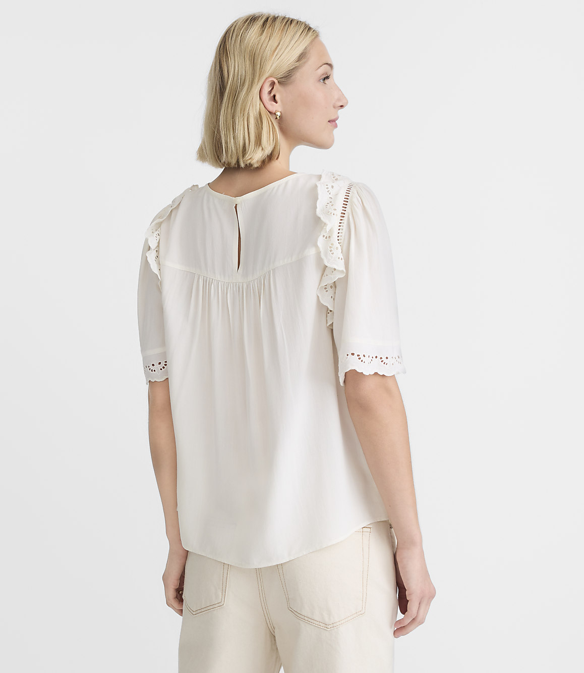 A woman in a white eyelet-trimmed blouse and cream pants stands against a plain white background.