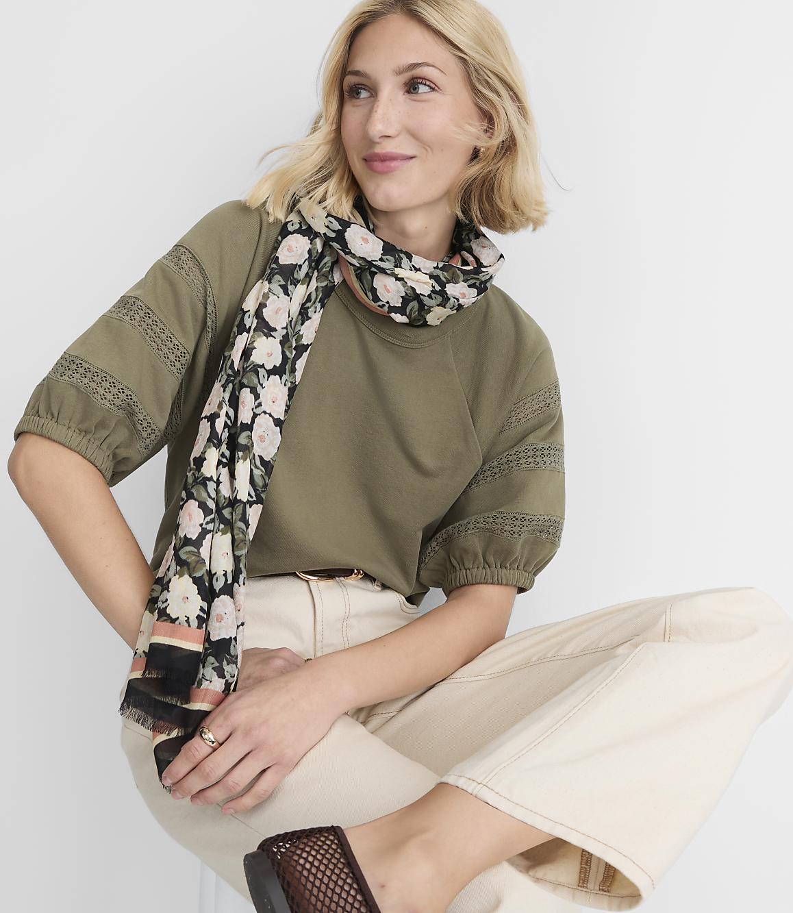 A blonde woman in a green top and cream pants, adorned with a floral scarf, sits against a plain white background.