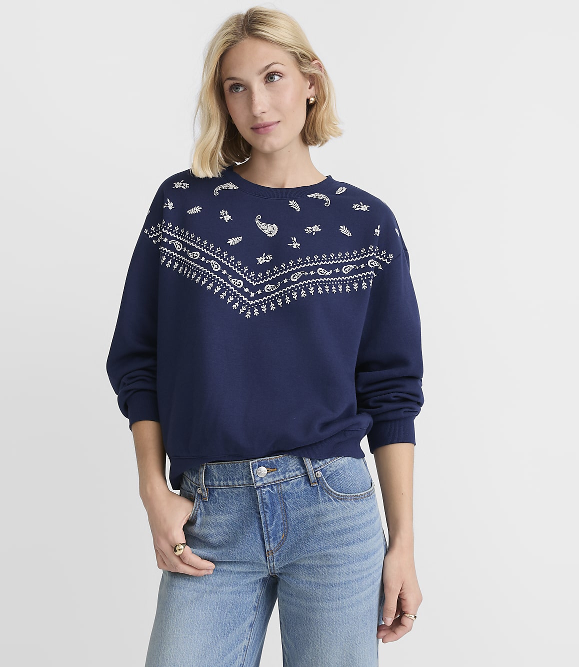 A woman in a navy sweatshirt with a white paisley pattern and blue jeans stands against a plain white background.