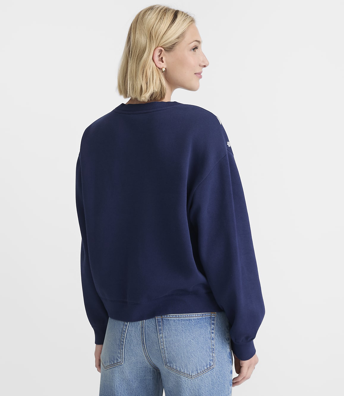 A woman in a navy sweatshirt and jeans stands against a plain white background.