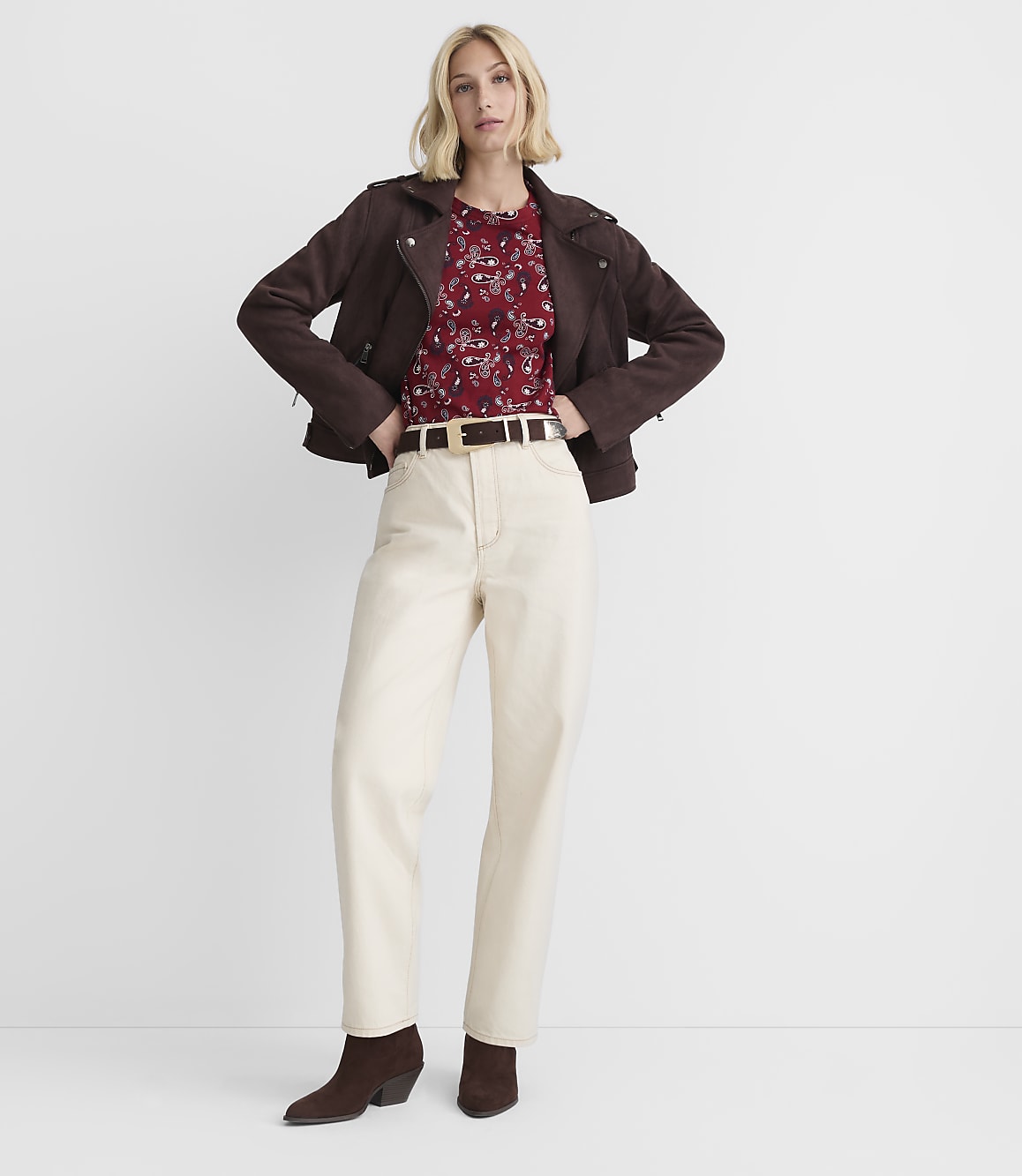 A woman in a dark brown jacket, red floral top, and cream pants stands against a plain white background.