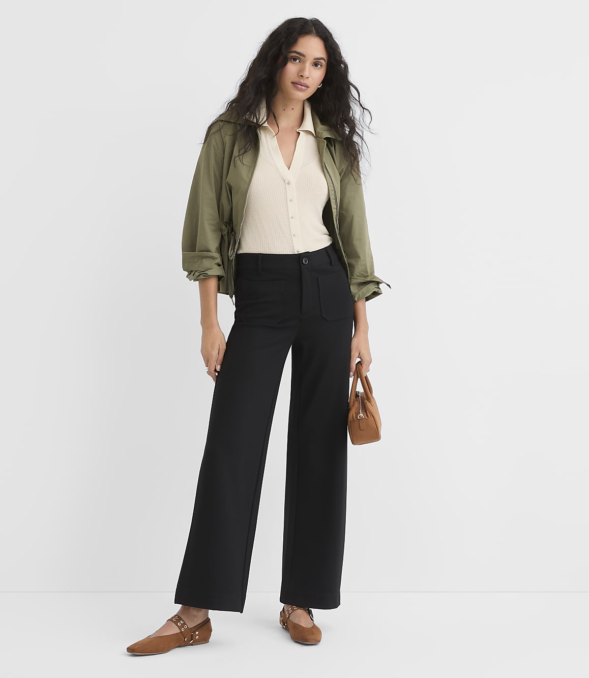A woman in a cream top, olive jacket, and black wide-leg pants stands against a plain white background, holding a small brown purse.