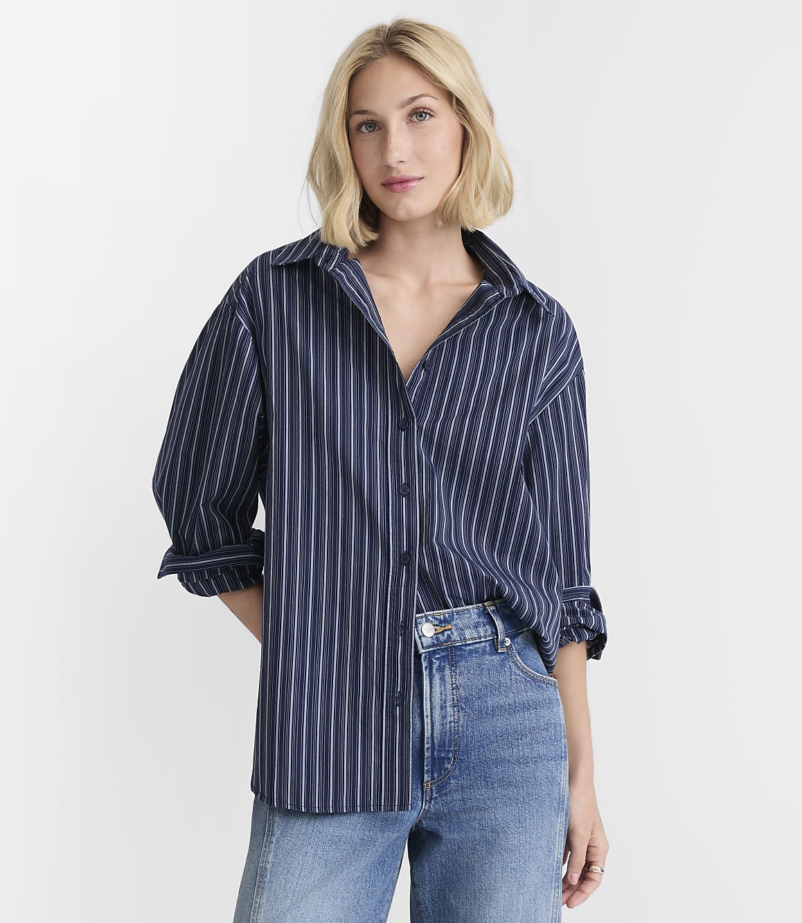 A blonde woman in a blue pinstripe shirt and jeans stands against a plain white background.