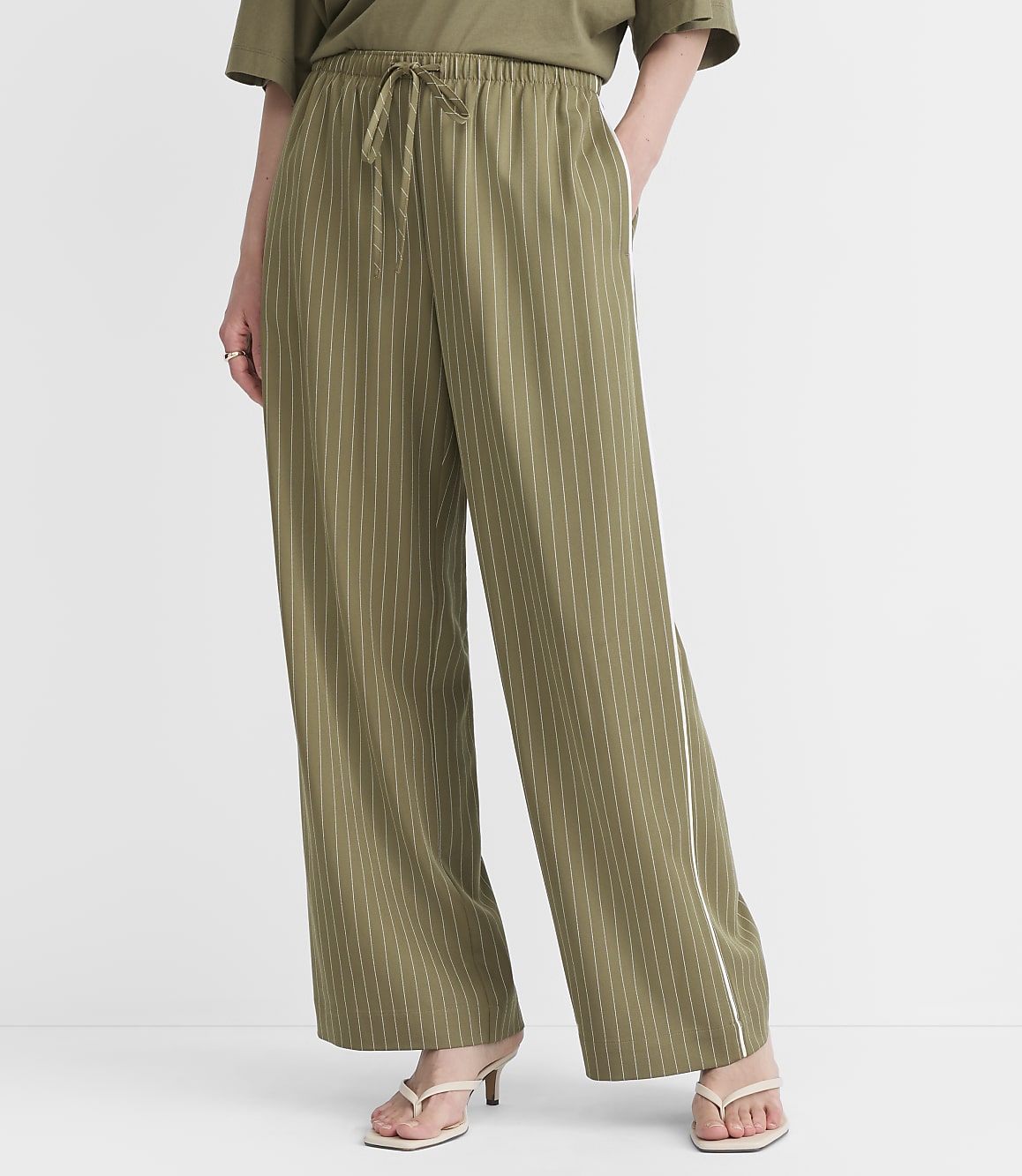 A person wearing olive green pinstripe pants and white sandals stands against a plain white background.