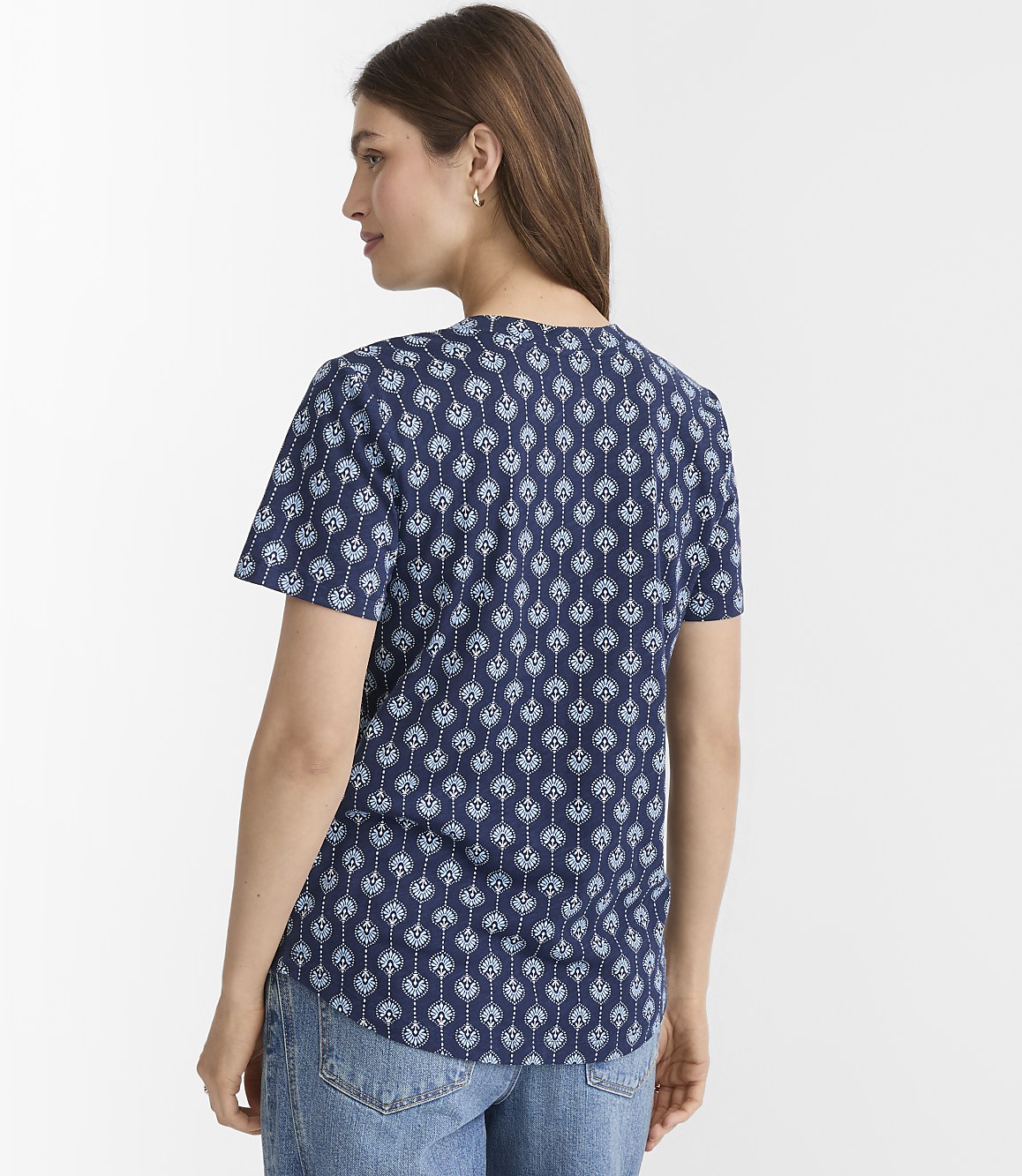 A woman in a patterned blue shirt and jeans stands against a plain white background.