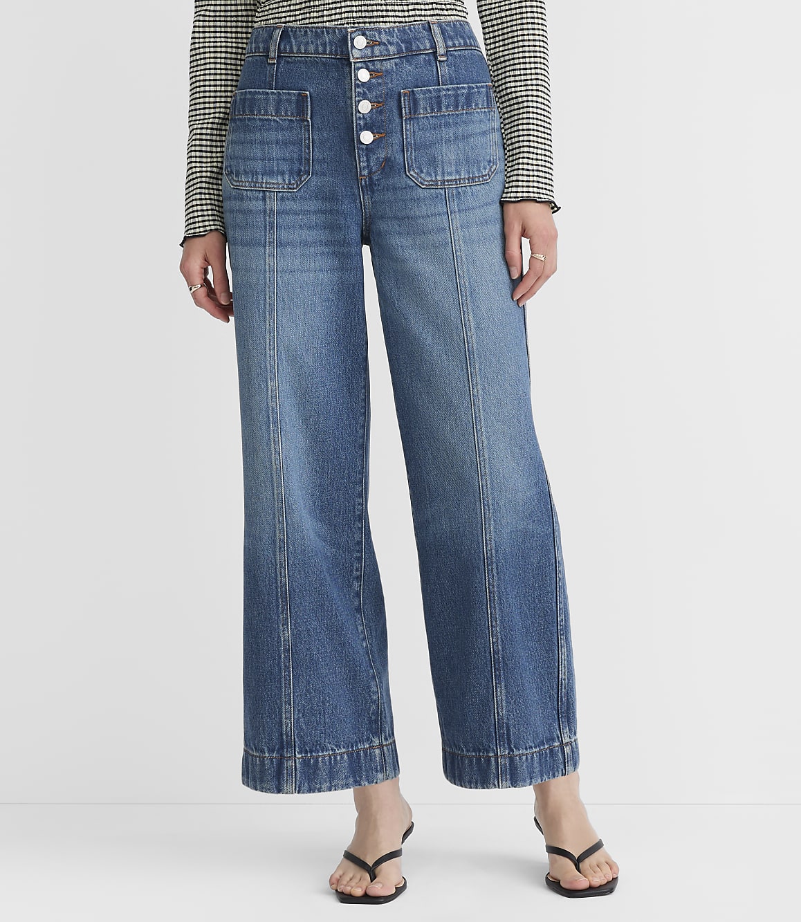 A person stands in front of a white background wearing wide-leg denim jeans with a button-up fly and a checkered long-sleeved shirt.