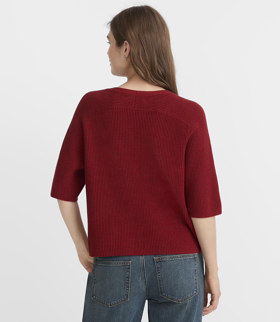 A woman in a red sweater and blue jeans stands against a plain white background.