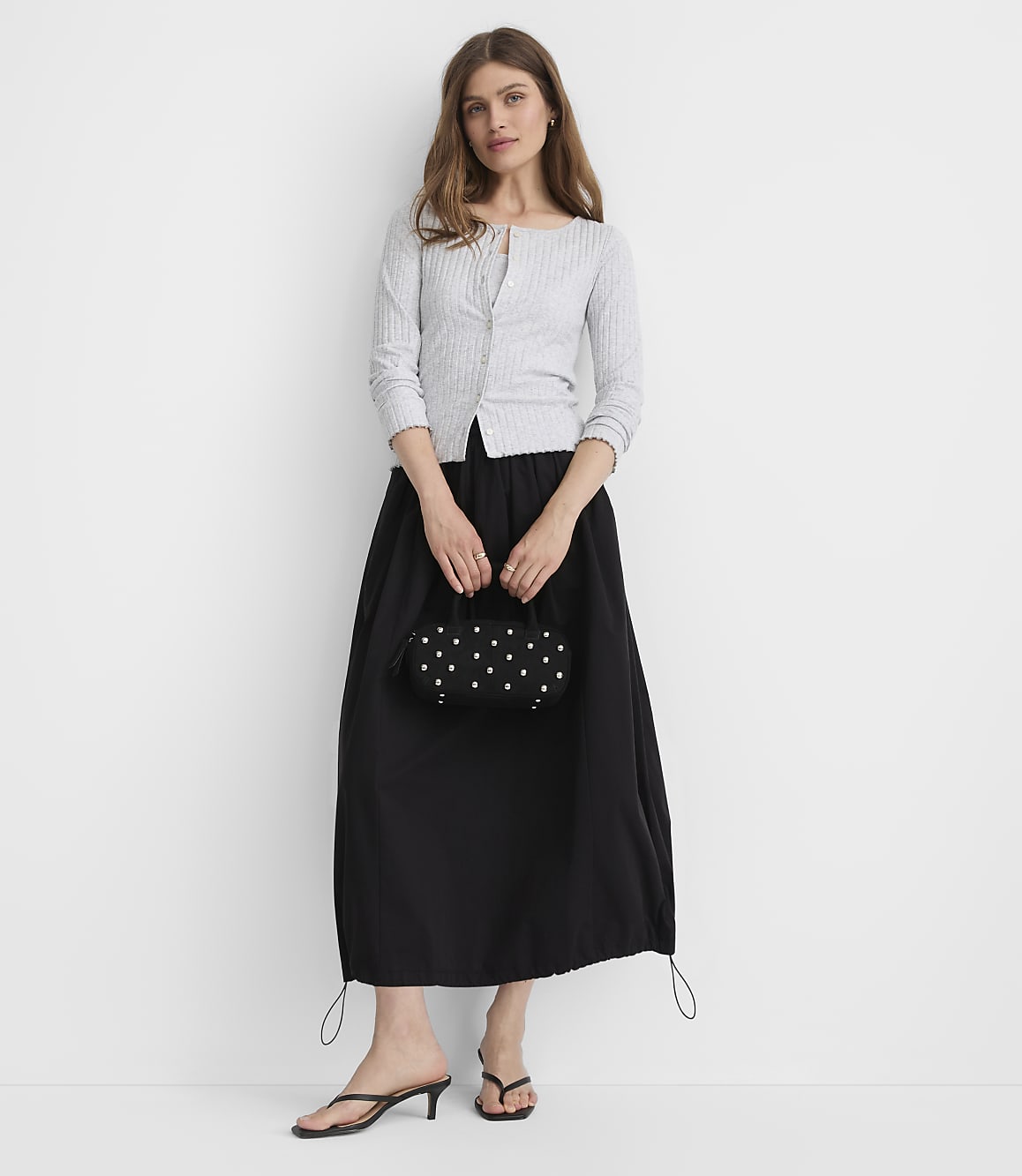 A woman in a light gray cardigan and black skirt stands against a plain white background, holding a small beaded purse.