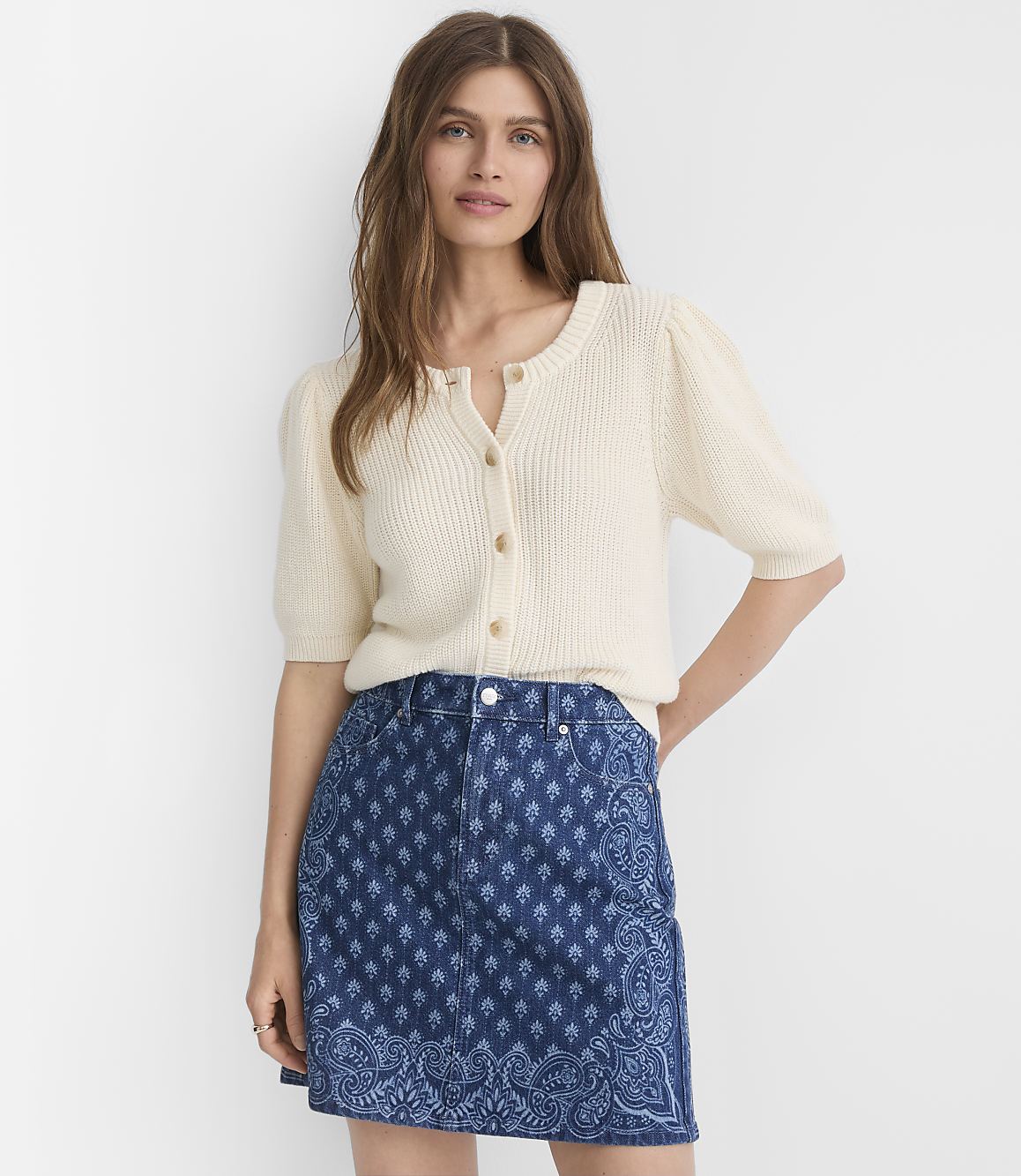 A woman in a cream-colored cardigan and a patterned denim skirt stands against a plain white background.