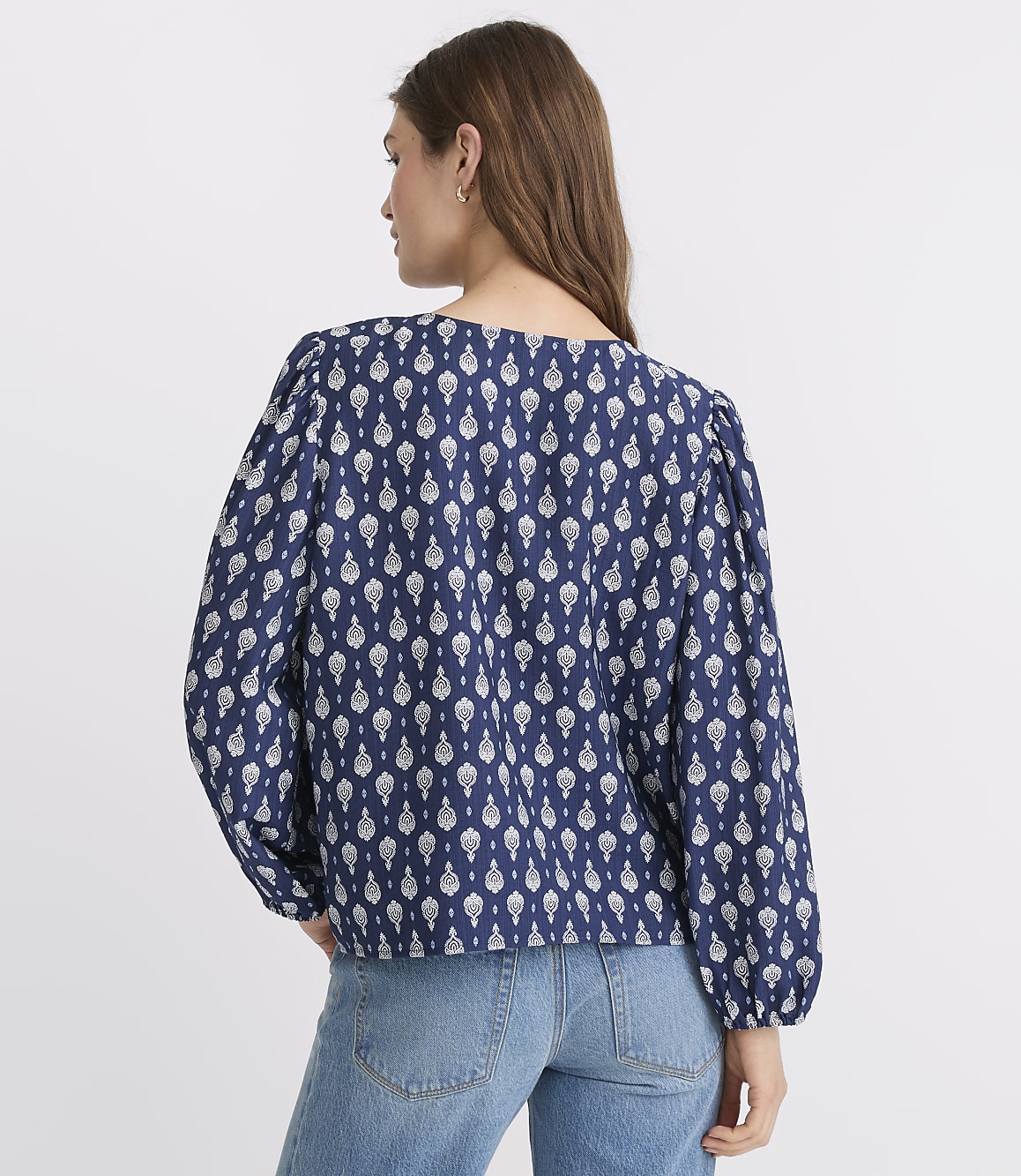 A woman in a blue and white patterned blouse and jeans stands against a plain white background.