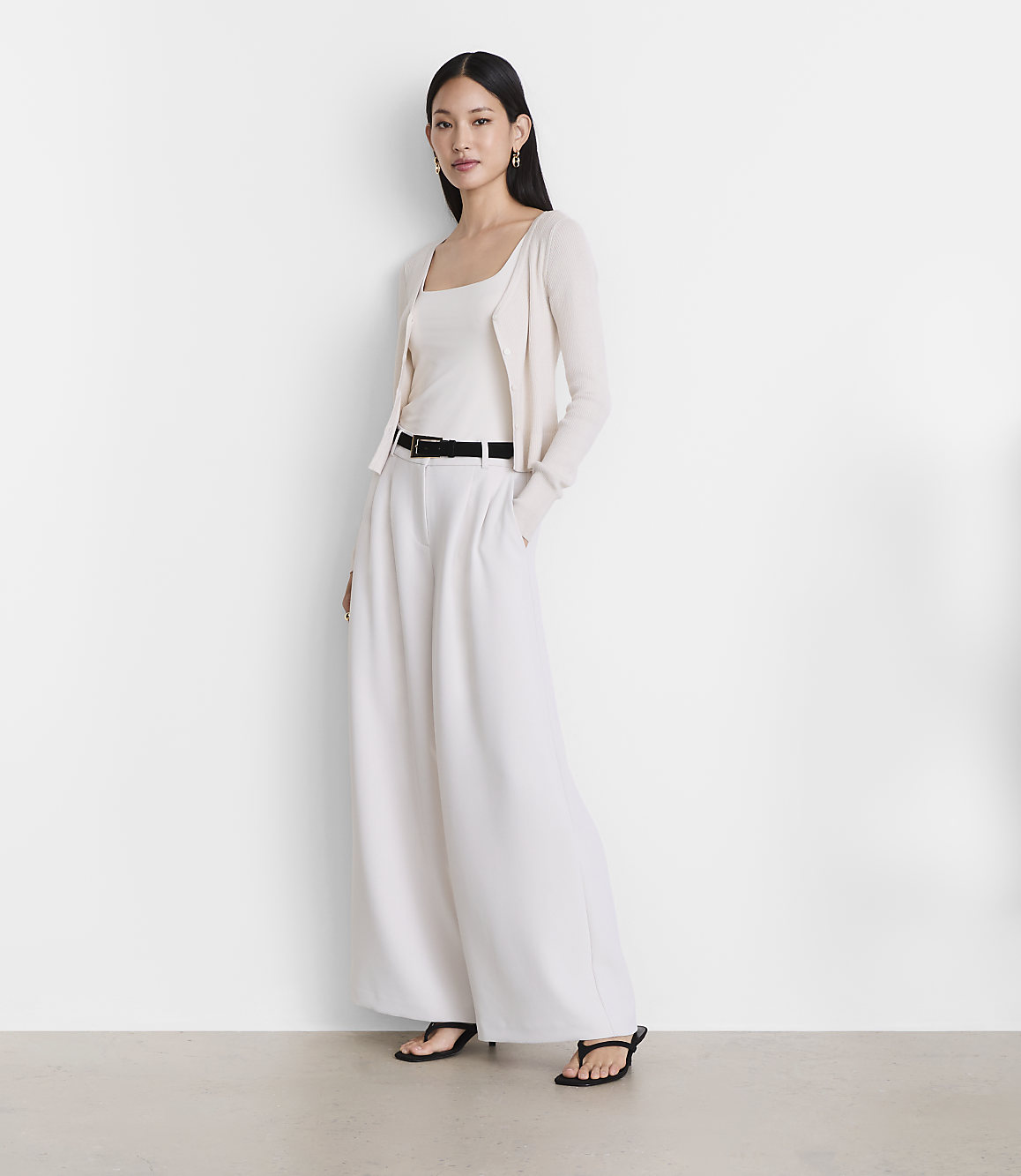 A woman in a cream-colored tank top, cardigan, and wide-leg pants stands against a plain white background.