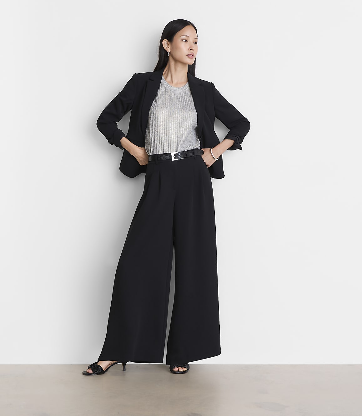 A woman in a black blazer and wide-leg pants stands against a plain white background.