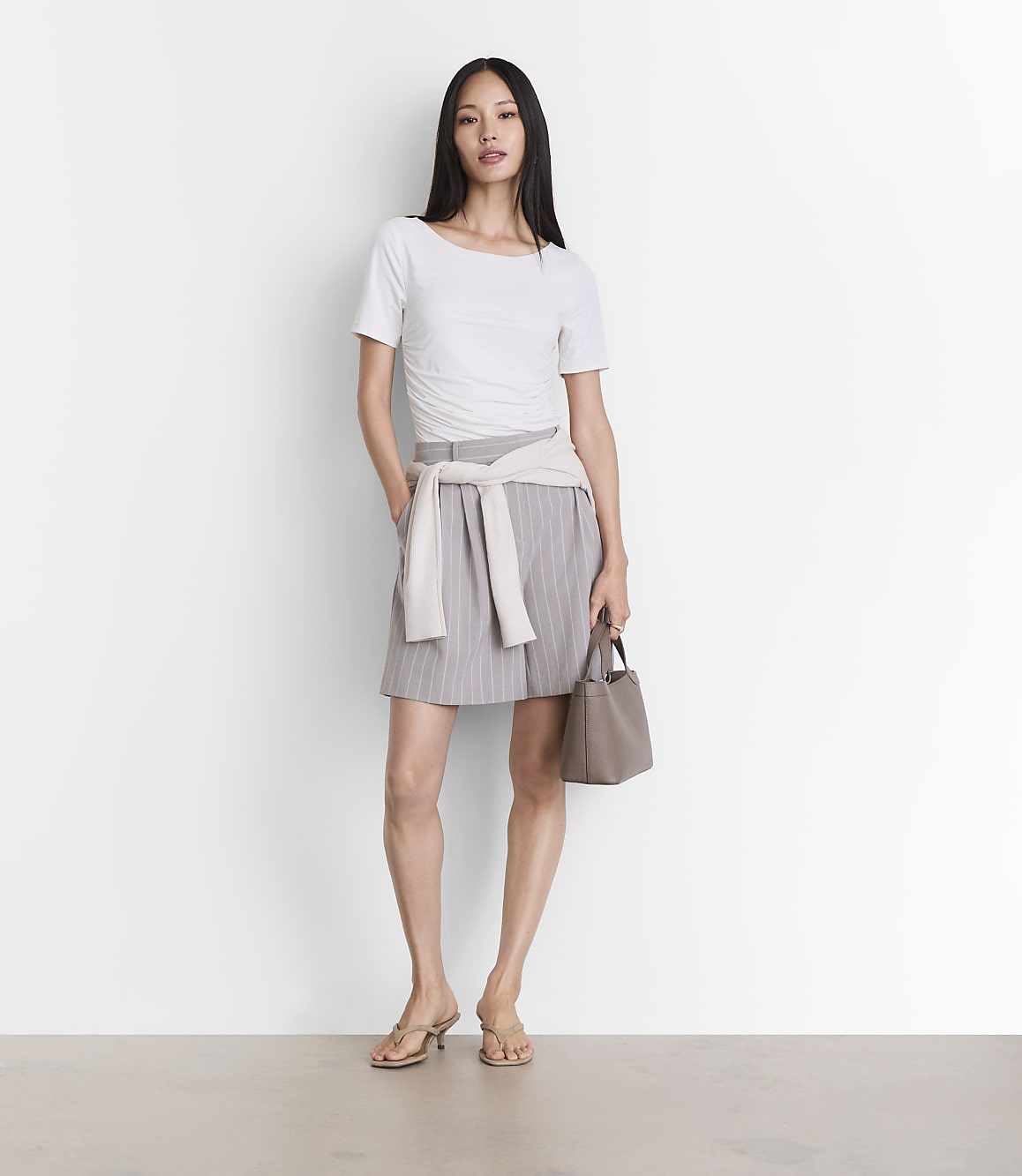 A woman in a white top and pinstripe skirt stands against a plain white background, holding a taupe handbag.