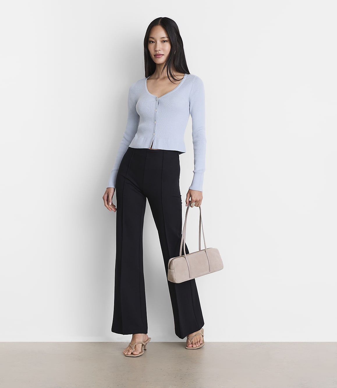A woman in a light blue cardigan and black flared pants stands with a beige handbag against a plain white background.