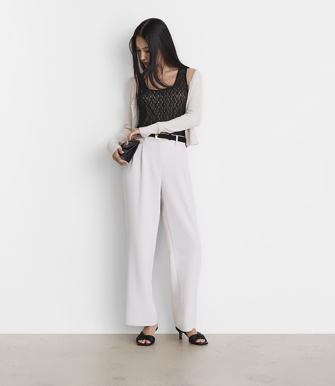 A woman in a black lace top, white pants, and a light cardigan stands against a plain white wall, holding a small black clutch.