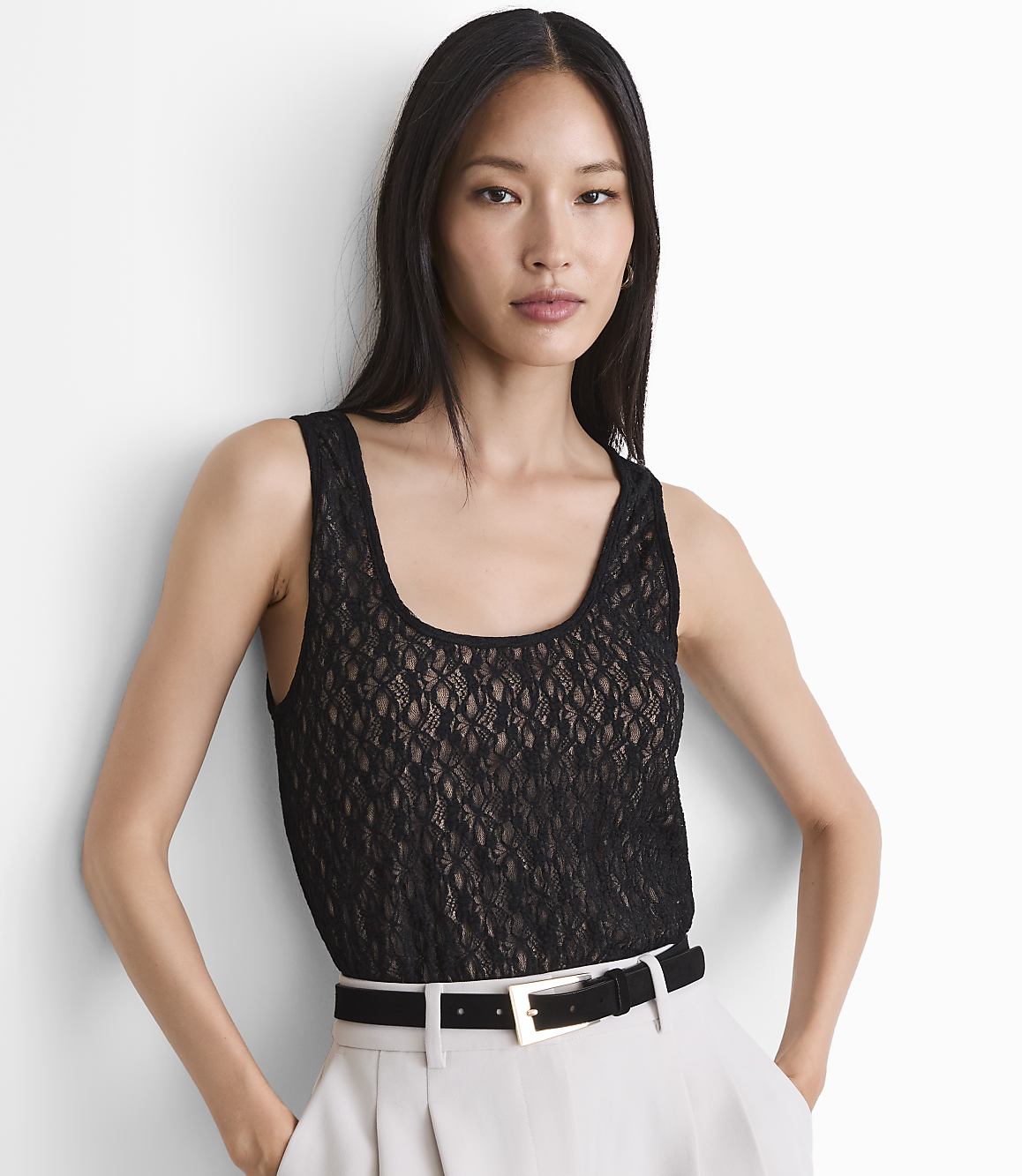 A woman in a black lace top and light-colored pants stands against a white background.