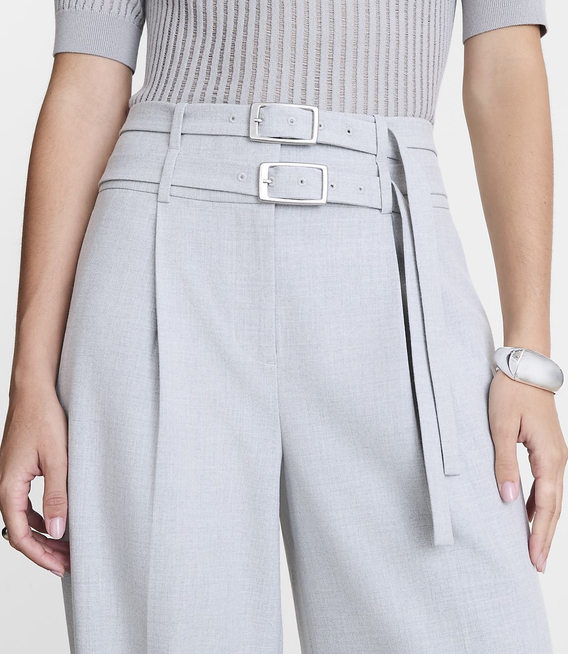 A person in a grey ribbed top and light grey pleated pants with a double buckle belt stands against a plain white background.