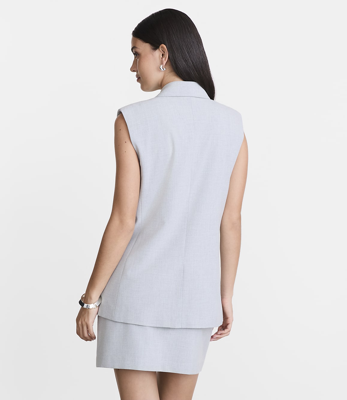 A woman in a light gray sleeveless blazer and skirt stands against a plain white background.
