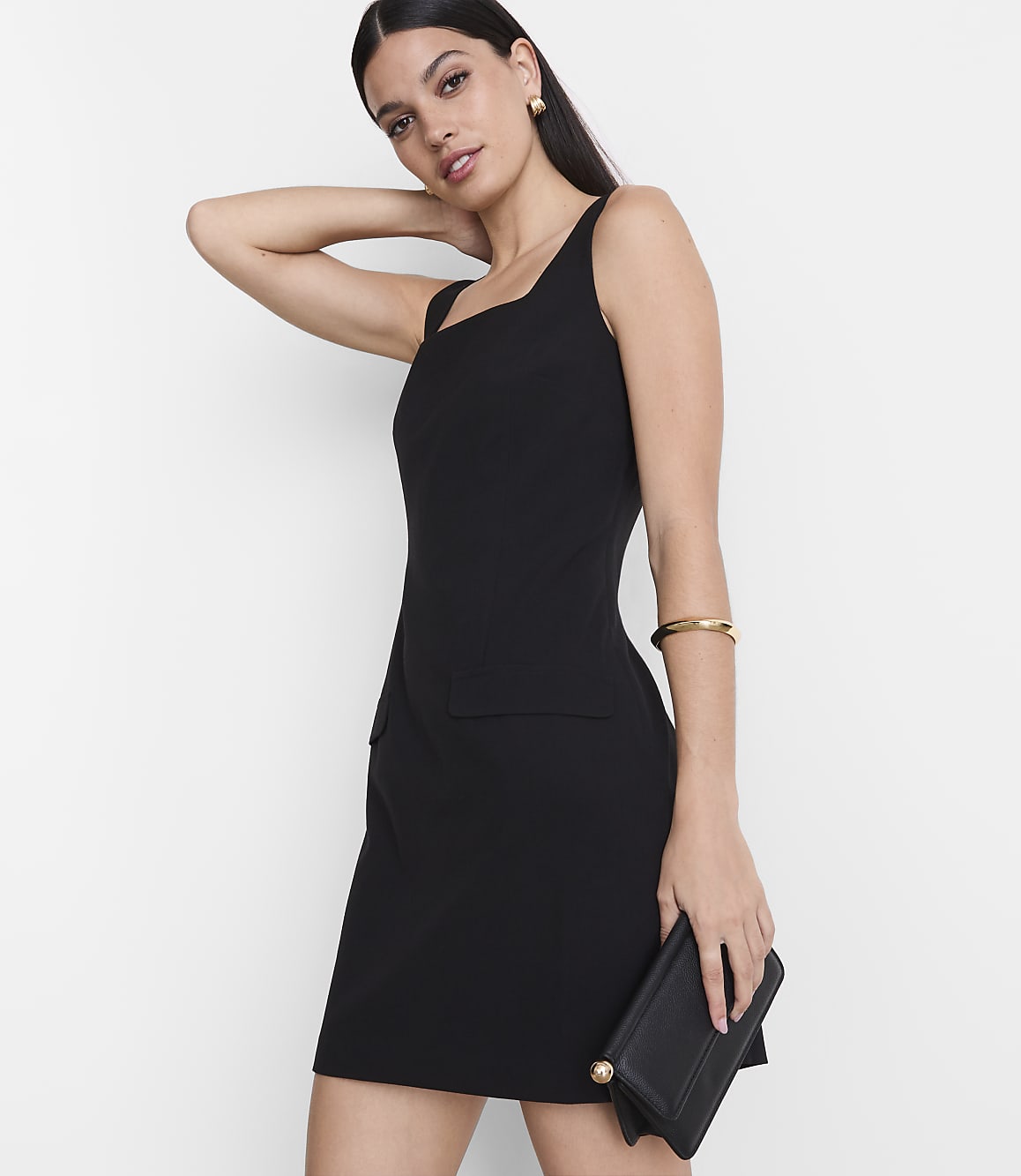 A woman in a black dress and holding a clutch stands against a plain white background.