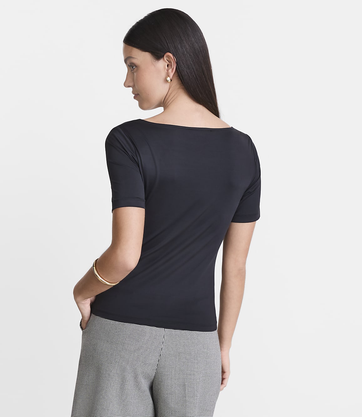 A woman in a black boatneck shirt and checkered pants stands against a plain white background.