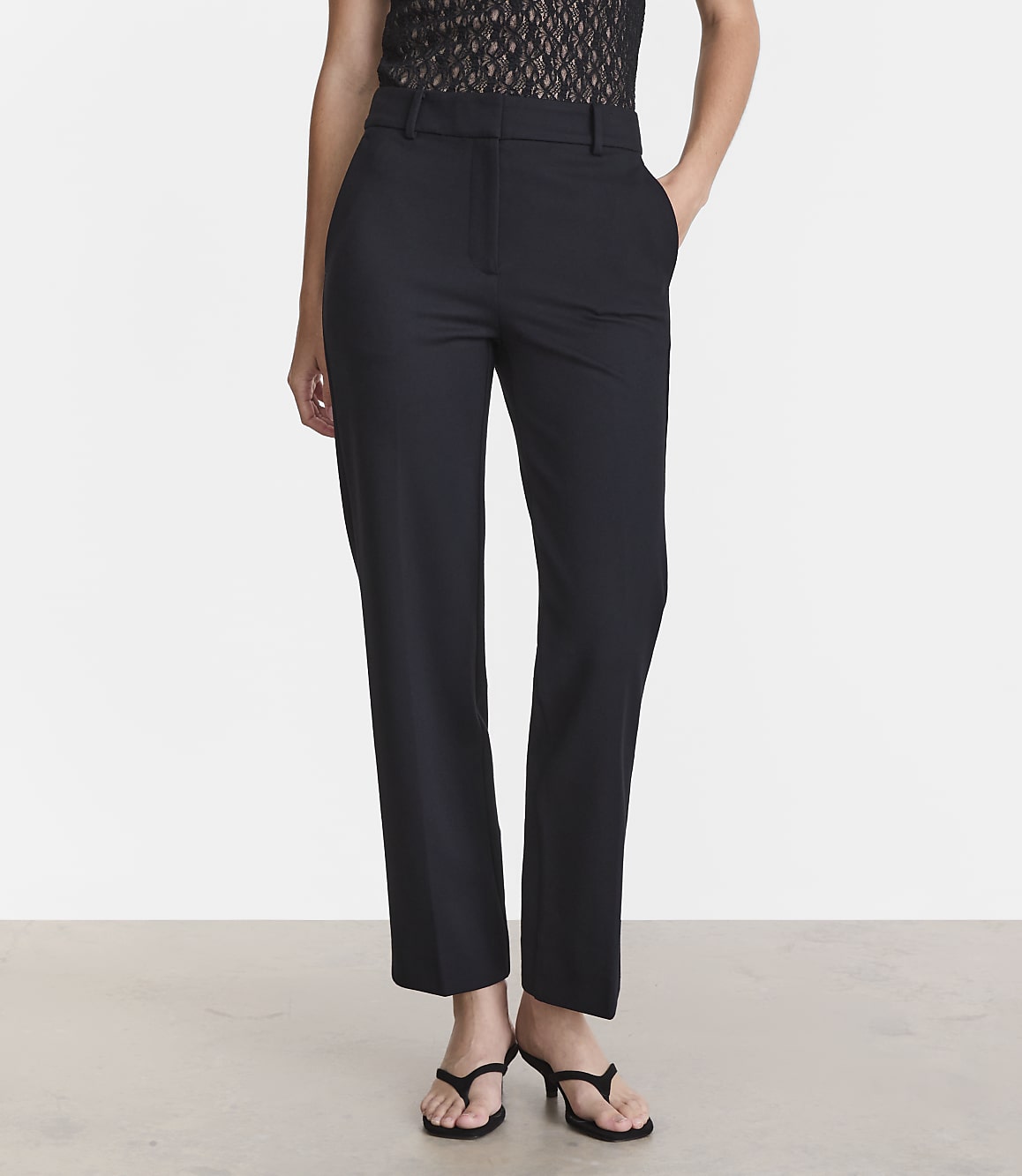 A person stands in black trousers and a lace top against a plain white background.