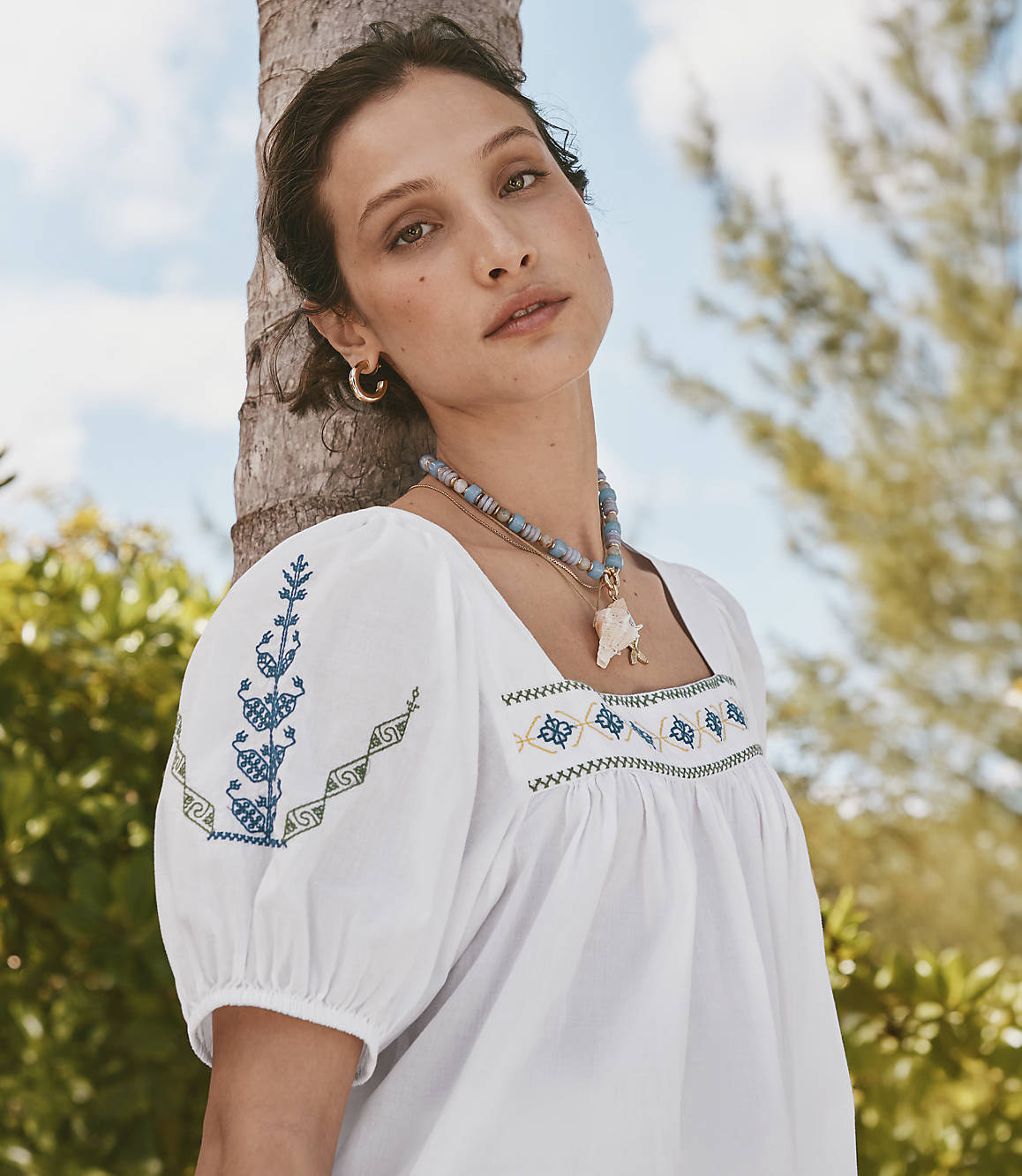 A woman with a beaded necklace and embroidered white top leans against a palm tree with a blurred tropical background.
