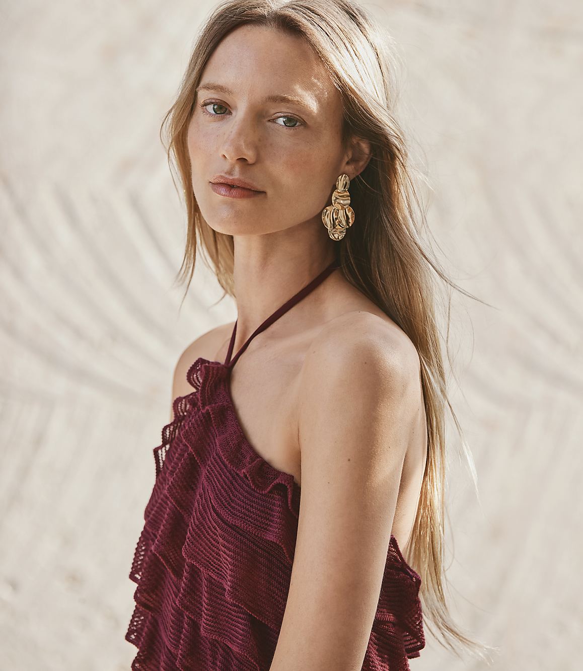 A woman with long blonde hair and large gold earrings wears a ruffled maroon halter top, with a sandy, palm-leaf-patterned background.