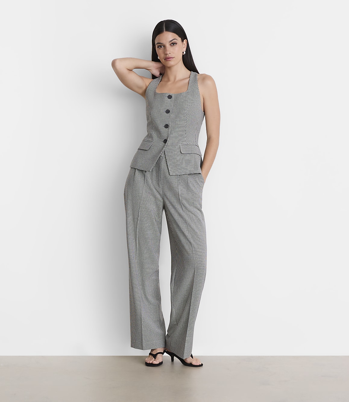 A woman in a checkered vest and pants stands against a plain white wall.