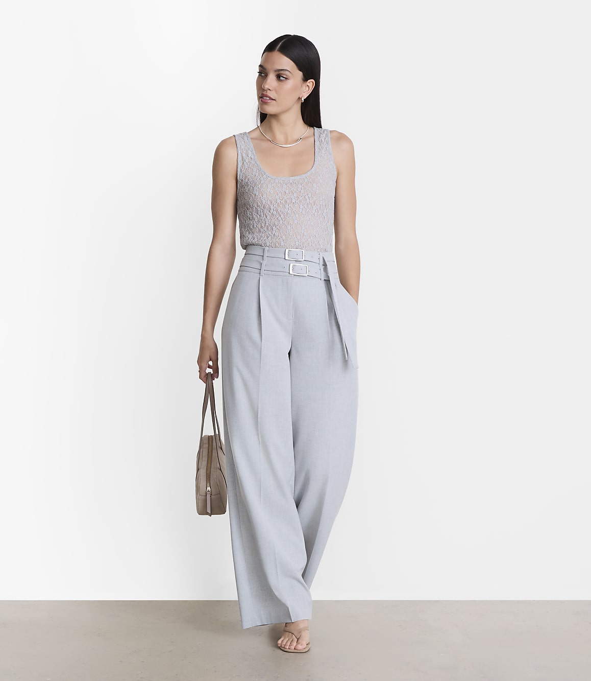 A woman in a light gray tank top and pale blue wide-leg pants stands against a plain white background, holding a tan tote bag.