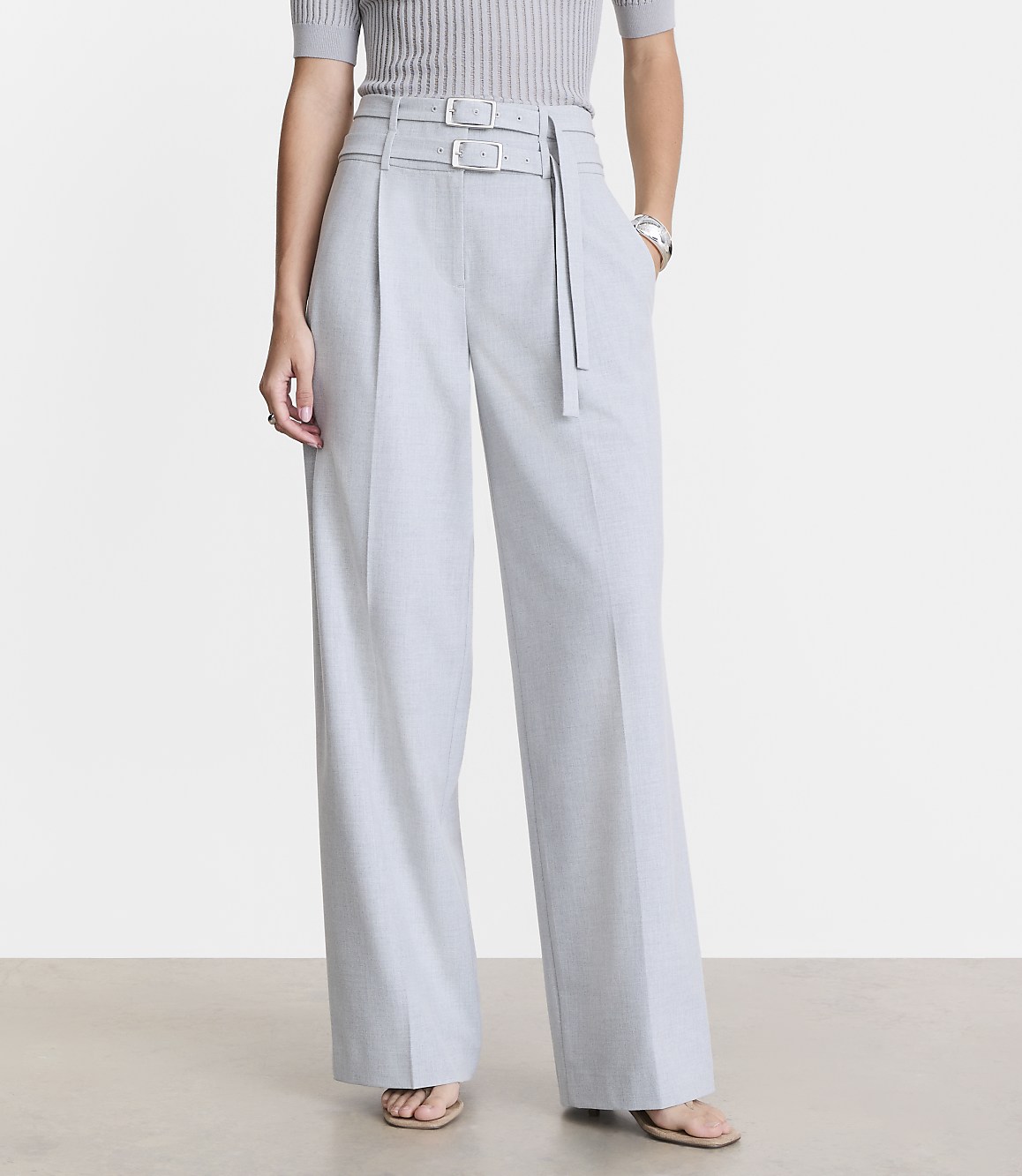 A person stands in light gray wide-leg pants with a double-buckle belt in the foreground, against a plain light gray background.
