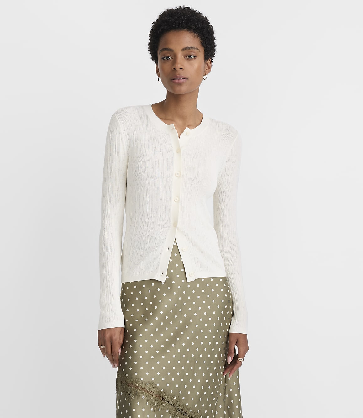 A woman in a cream-colored cardigan and a polka-dot skirt stands against a plain white background.