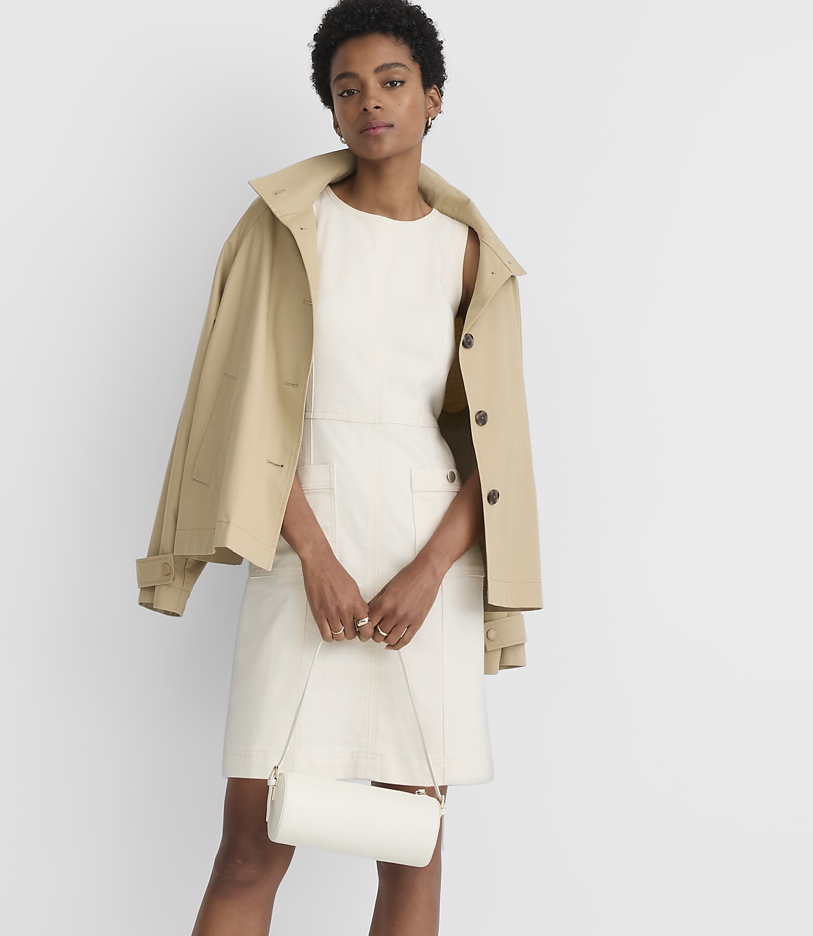 A woman in a cream dress and tan jacket holds a white cylindrical purse in front of a plain white background.