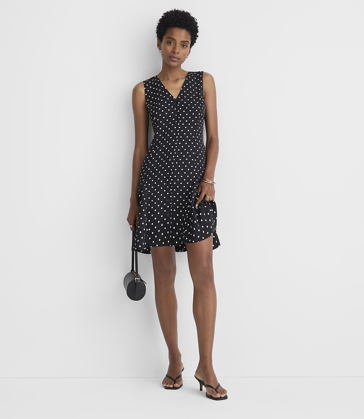 A woman in a polka dot dress and heels stands against a plain white background, holding a small black purse.