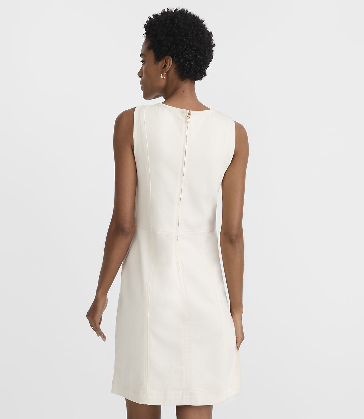 A woman in a cream-colored sleeveless dress stands against a plain white background.