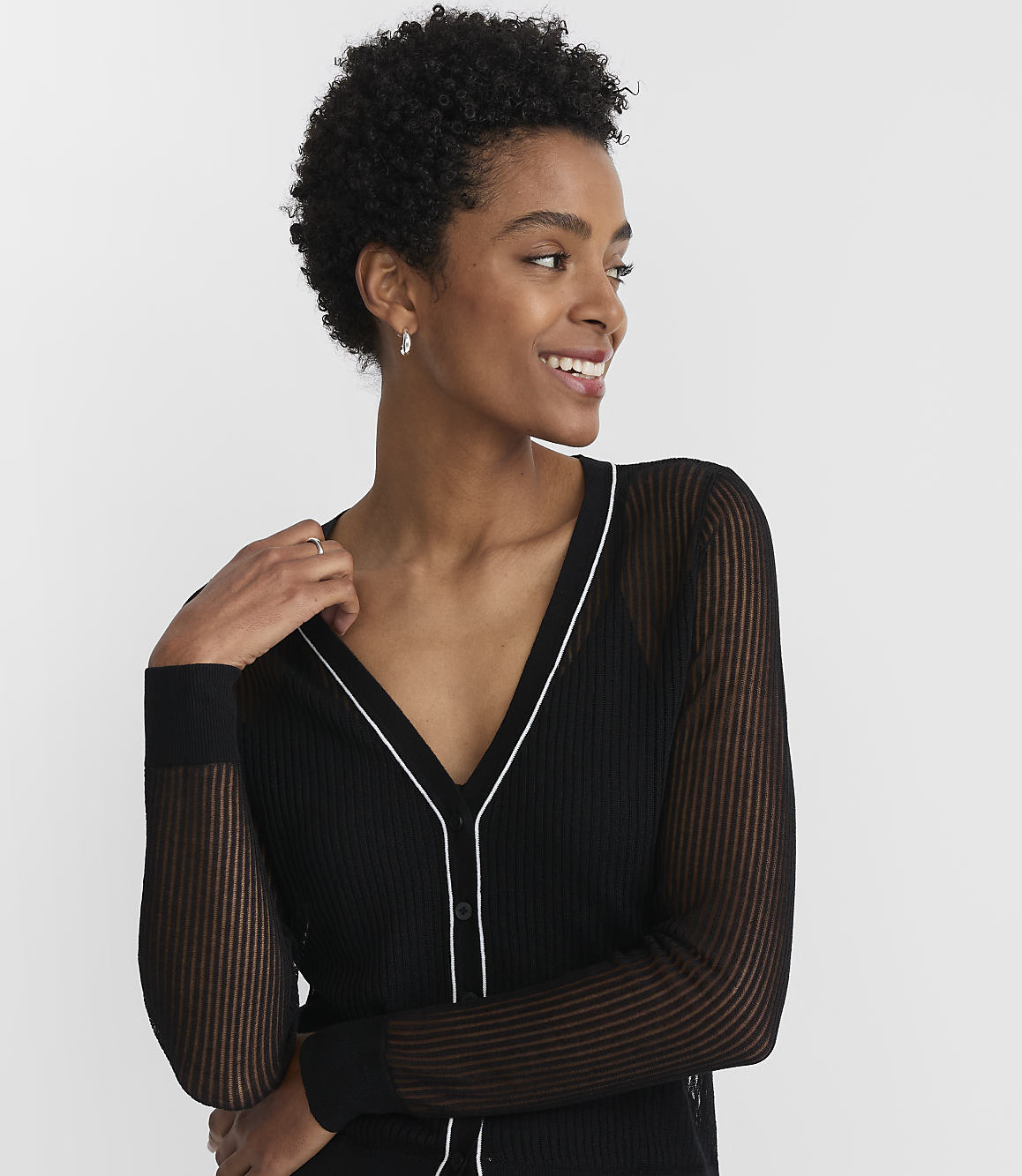 A woman with short, dark, curly hair smiles as she wears a black cardigan with sheer sleeves against a plain white background.