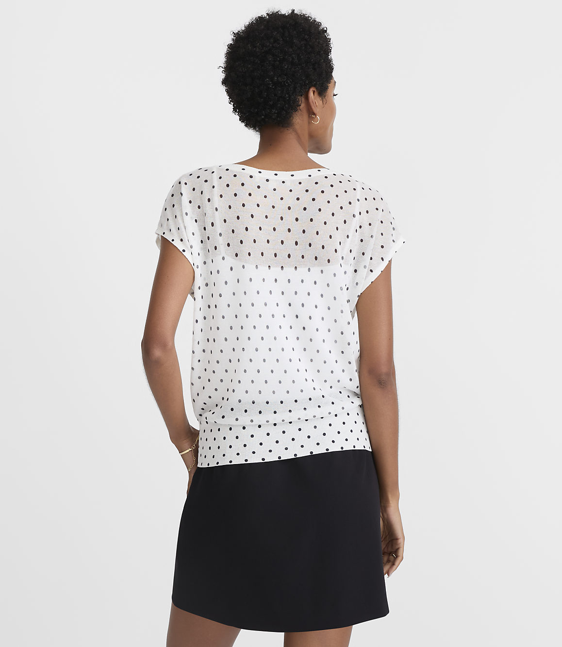A woman in a white polka-dot blouse and black skirt stands against a plain white background.