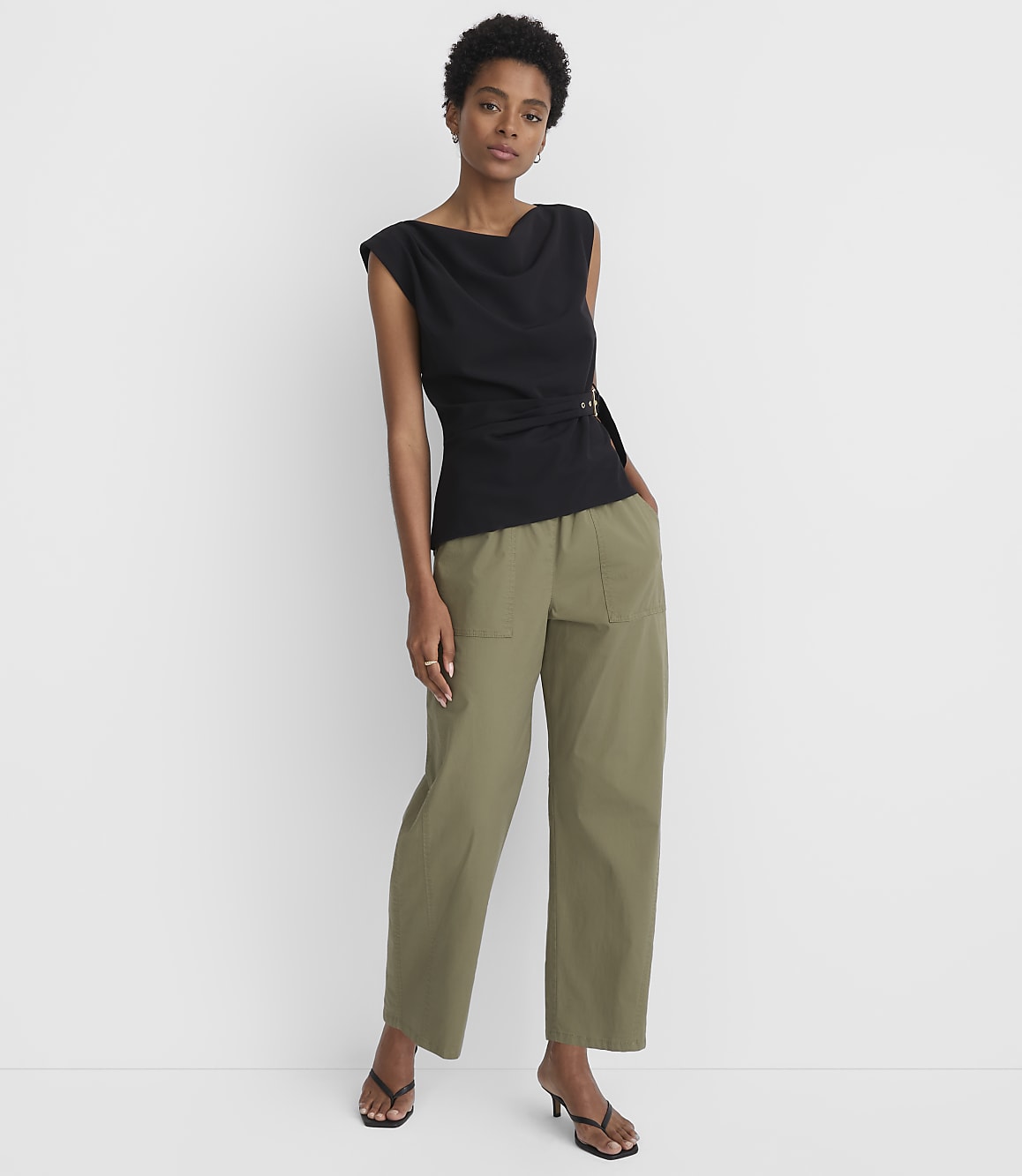 A woman in a black top and olive green pants stands against a plain white background.