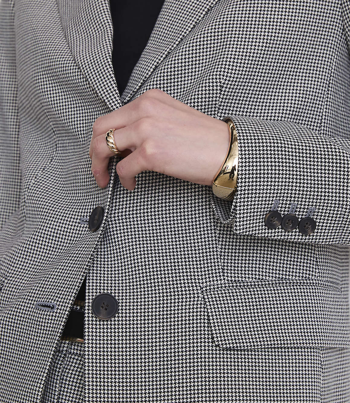 A person in a houndstooth suit adjusts their gold bracelet and ring, with a blurred background.
