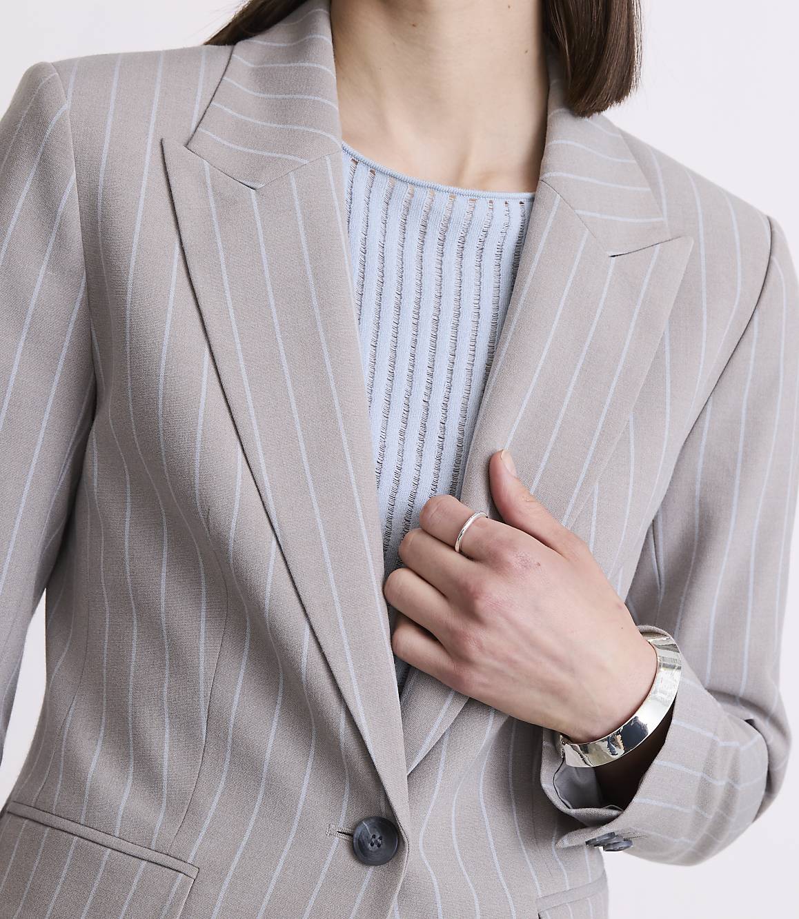 A woman in a pinstripe blazer and light blue top poses with her hand on her chest, against a plain background.