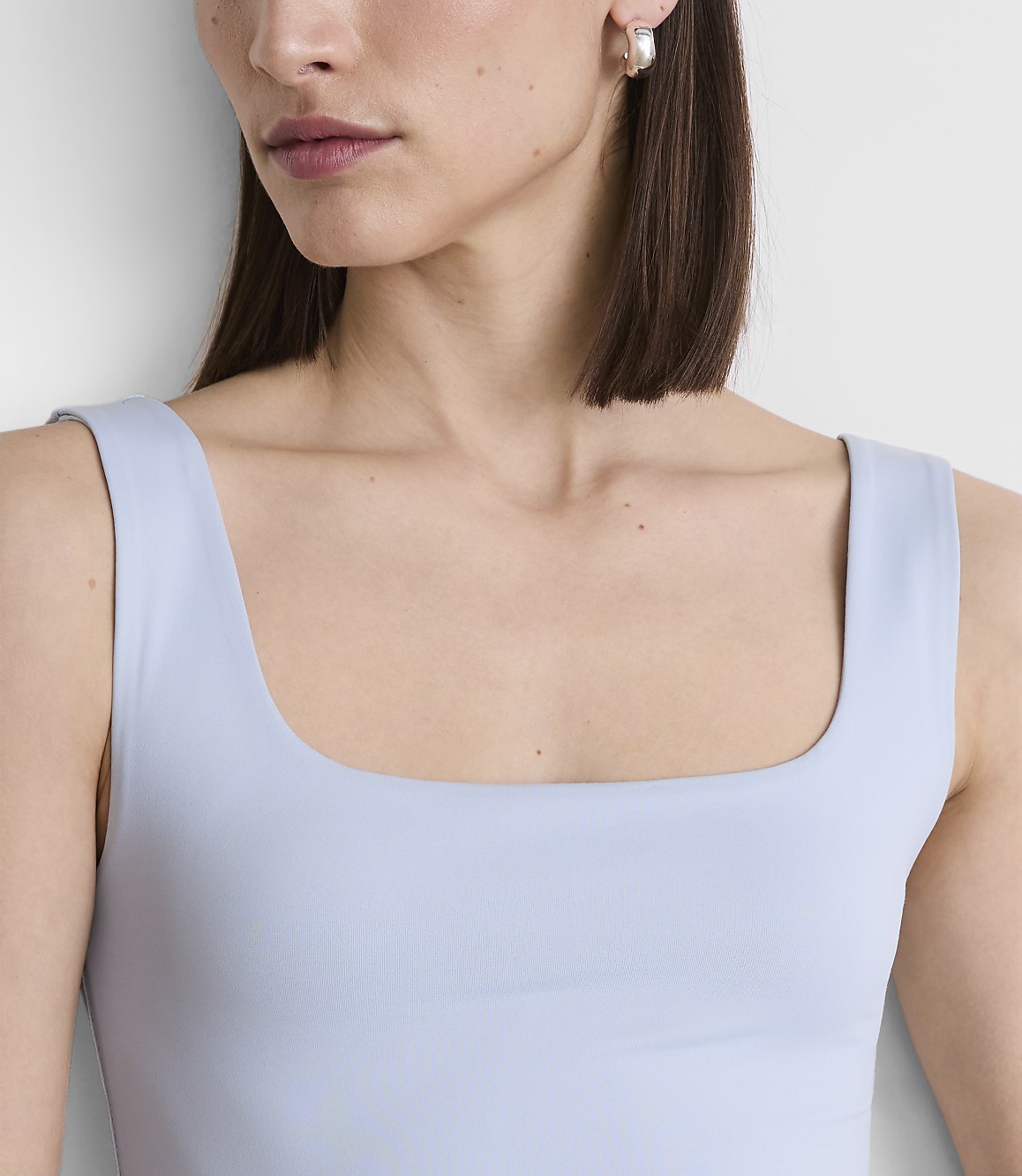 A woman with a square neckline top and a pearl earring is shown against a plain white background.