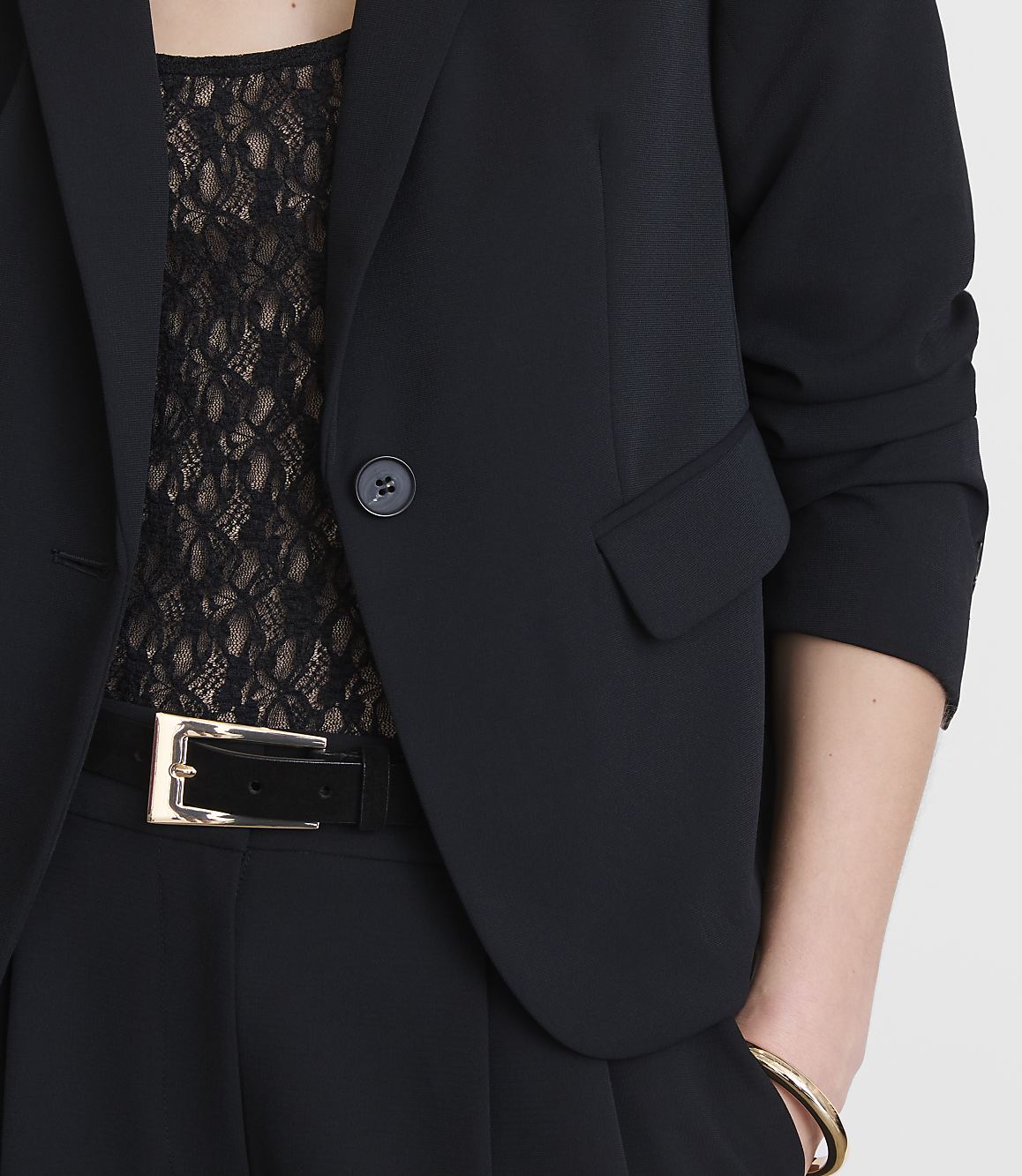 A woman in a black blazer and lace top, with a black belt and gold bracelet, stands against a plain white background.