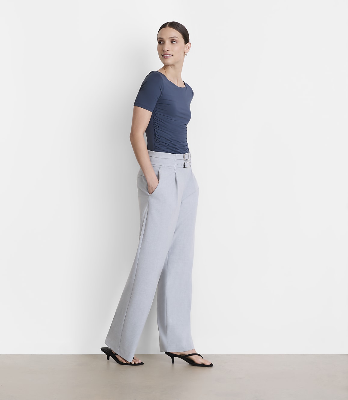 A woman in a blue ruched top and light gray wide-leg pants stands against a plain white background.