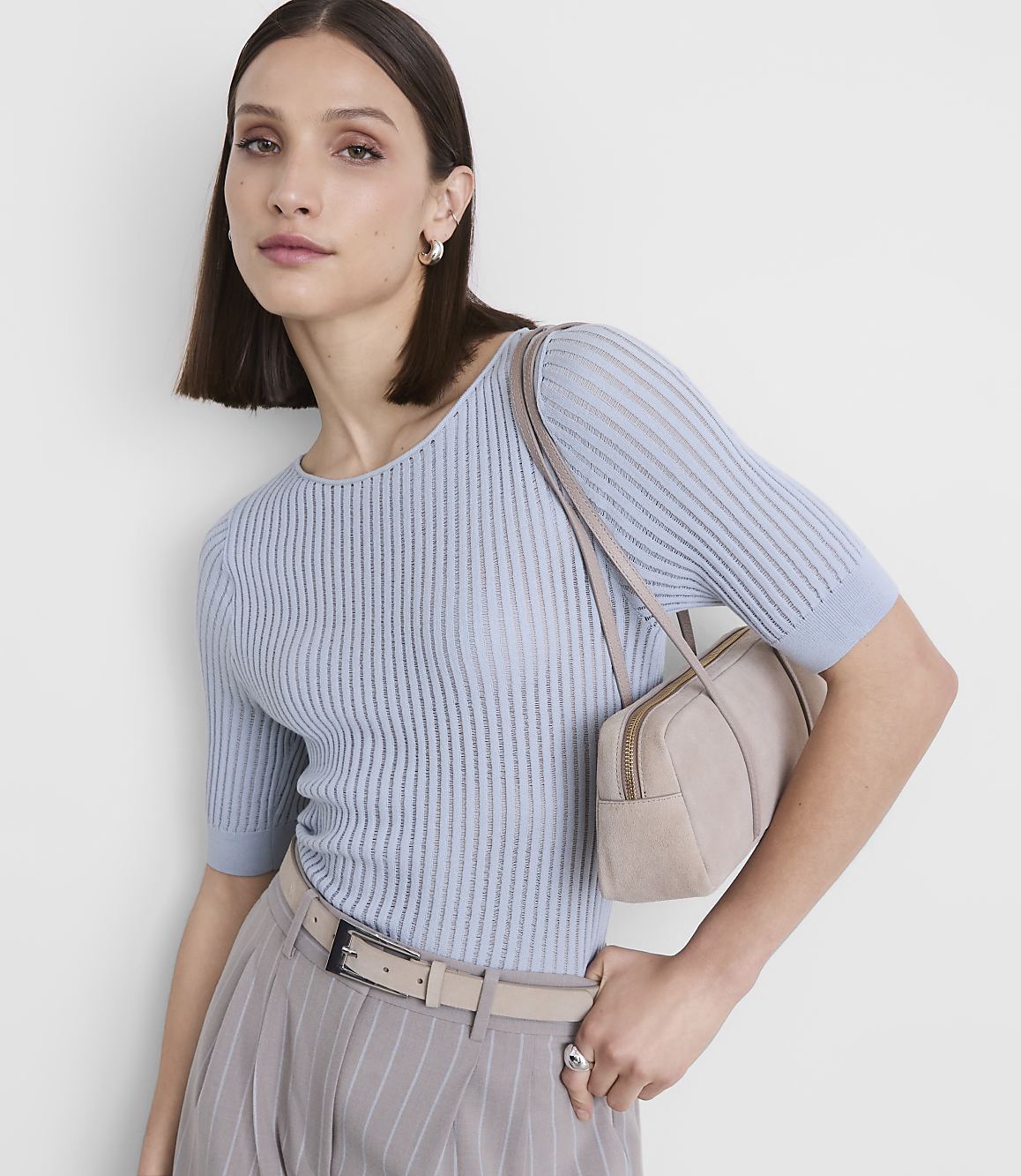 A woman in a light blue ribbed top and pinstripe pants poses with a small beige handbag against a plain white background.