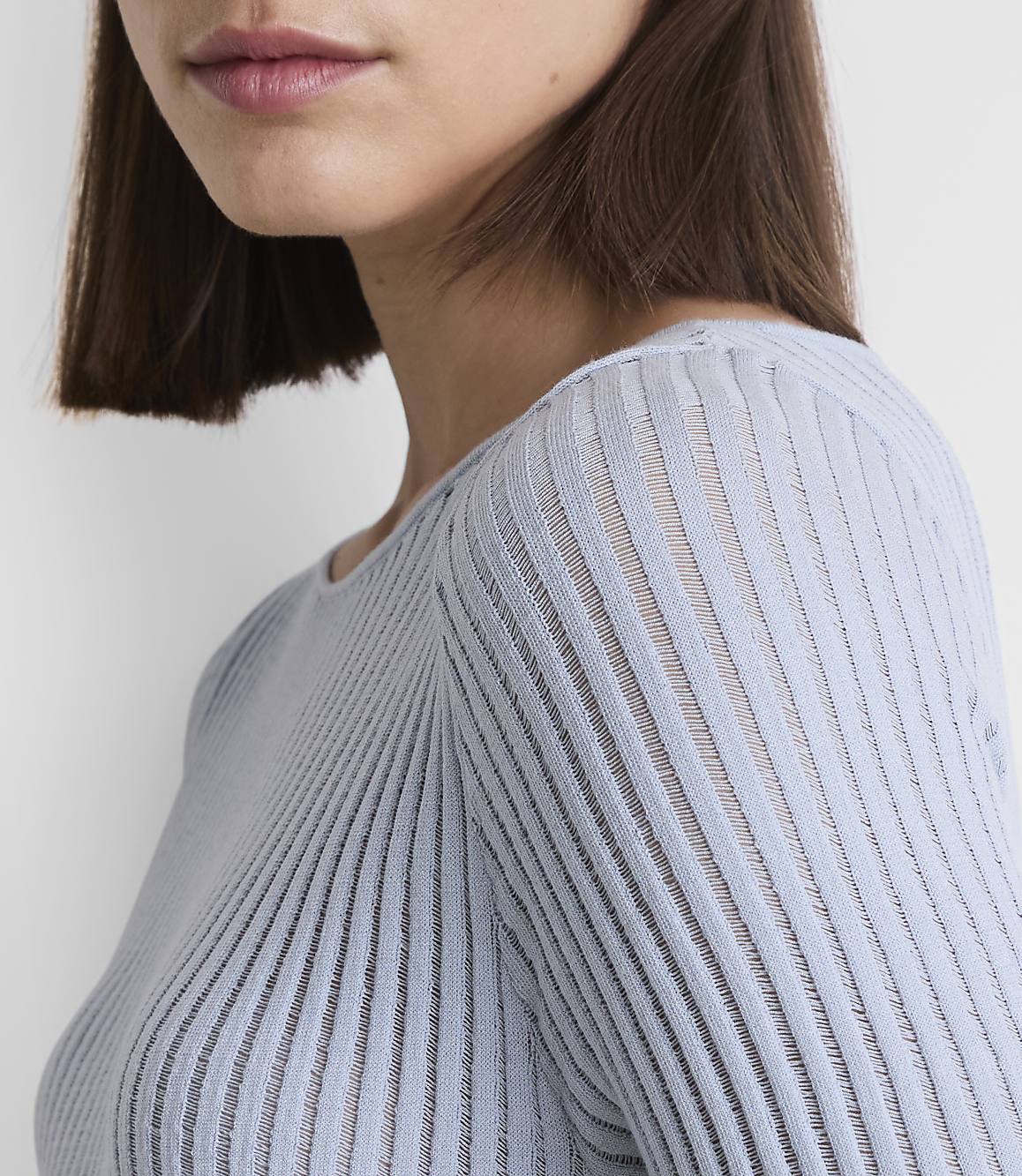 A close-up of a woman's shoulder and neck in a light blue ribbed sweater is set against a plain white background.